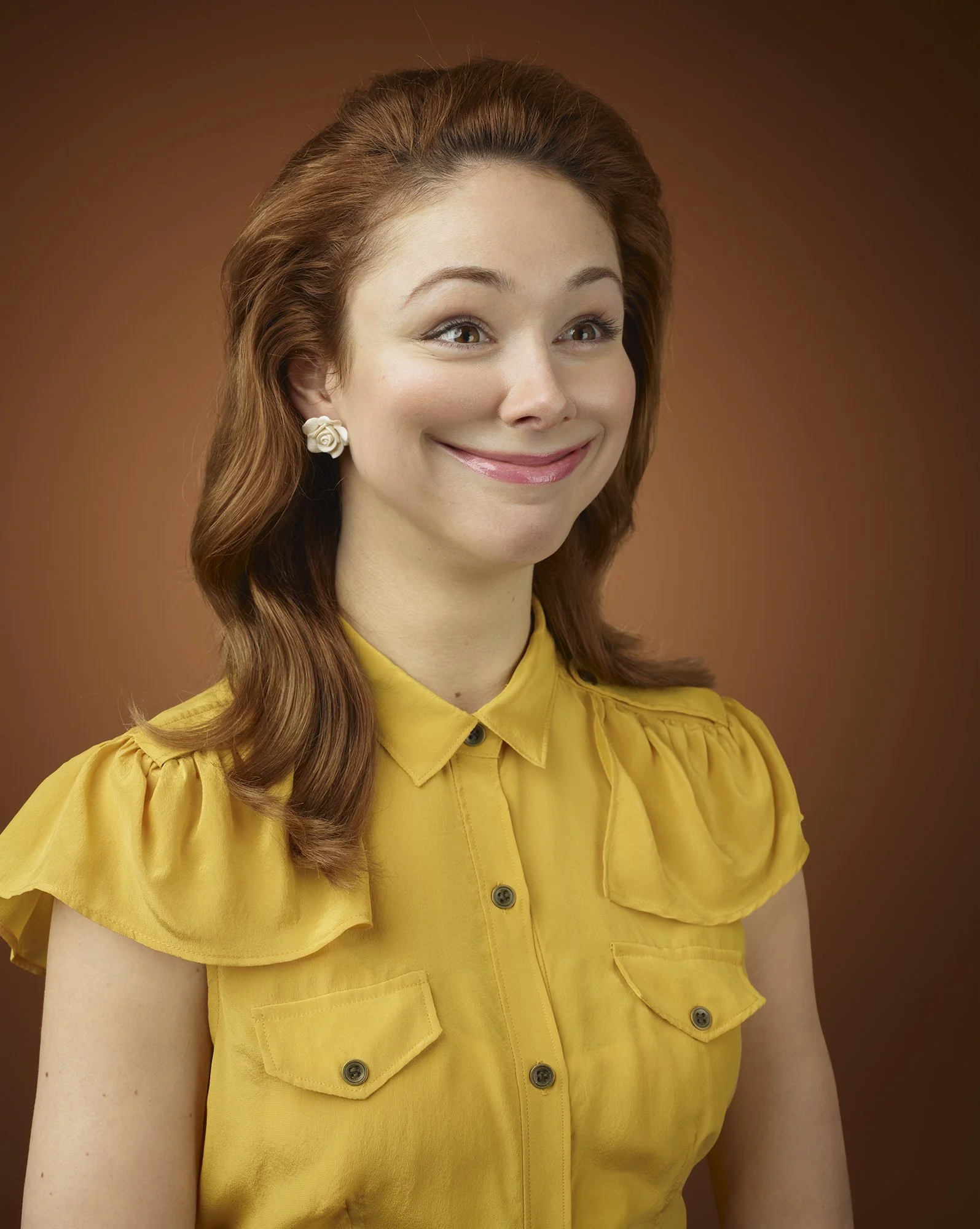 A woman with shoulder-length red hair smiling, wearing a yellow blouse with ruffled sleeves and white flower earrings, in front of a brown gradient background.