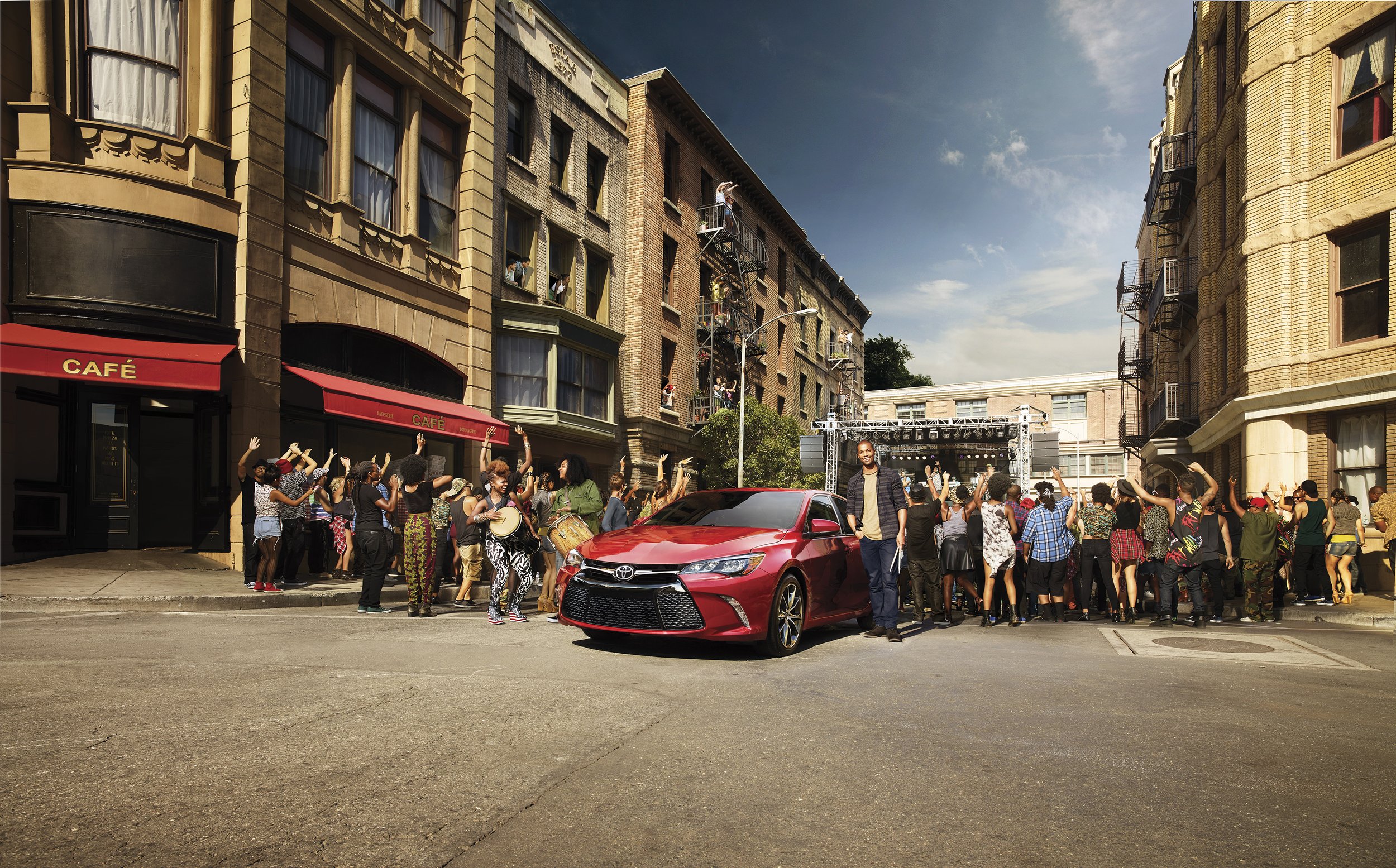 A lively city street scene with a crowd of people dancing and celebrating in front of a stage, a red car parked in the foreground, and tall brick buildings with fire escapes lining the street under a partly cloudy sky.