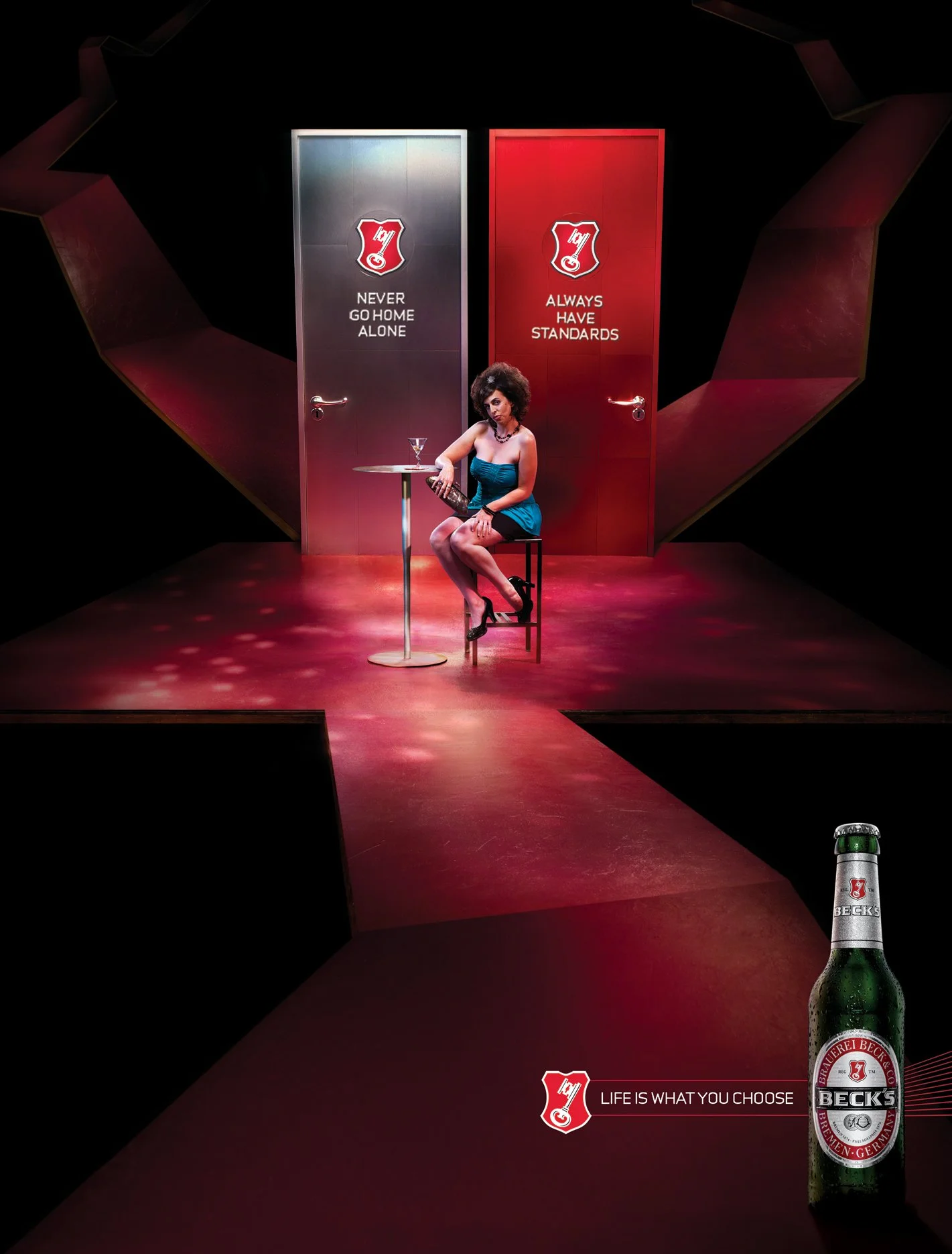 A woman sitting on a stool in a dimly lit room with red and black decor. Behind her are two doors with slogans: "Never Go Home Alone" and "Always Have Standards." A table with a drink and a Beck's beer bottle in the foreground displays the slogan "Li