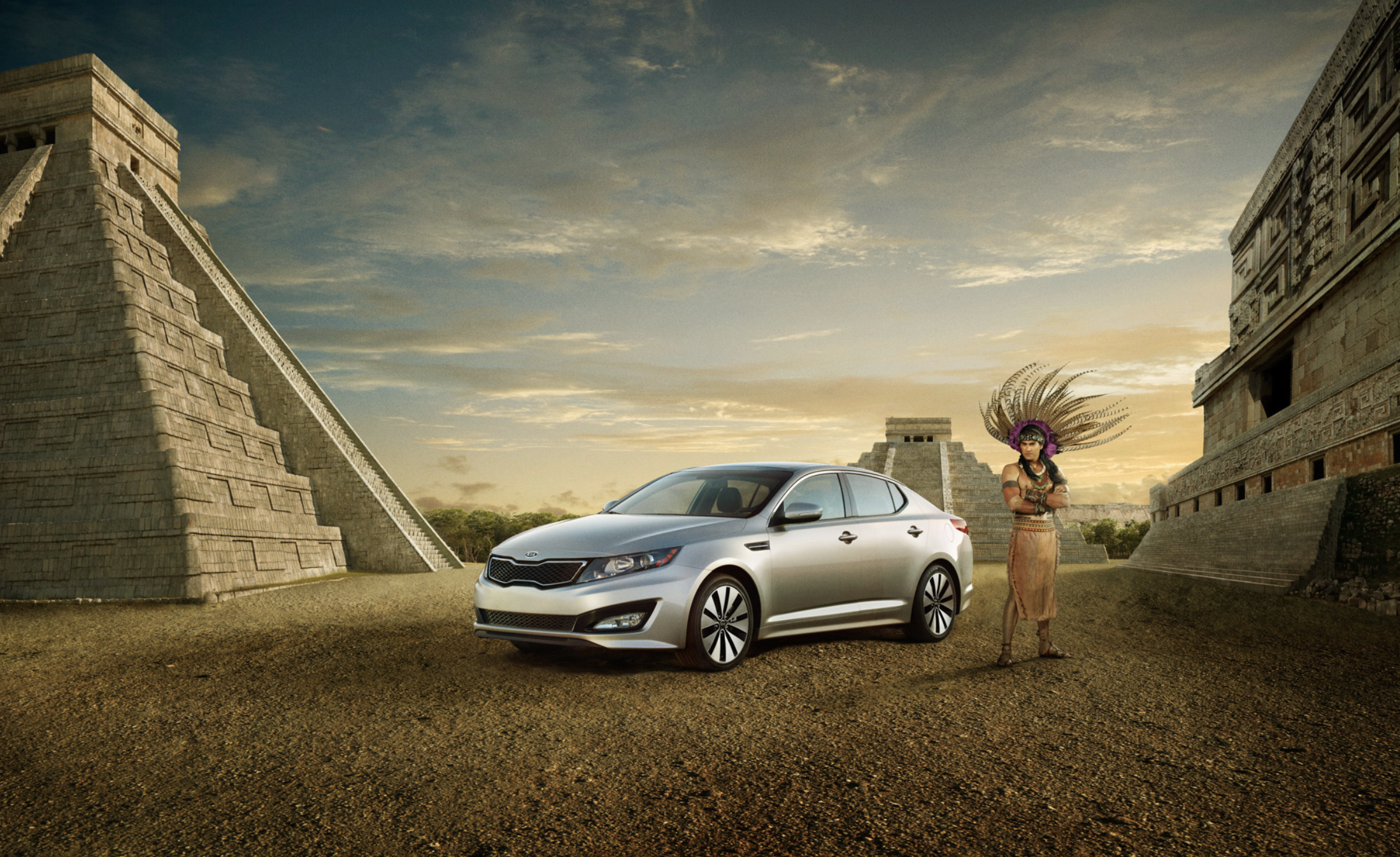 A silver Kia sedan parked on a dirt ground in front of ancient Mesoamerican pyramids at sunset, with a woman dressed in traditional indigenous attire and a feather headdress standing next to it.