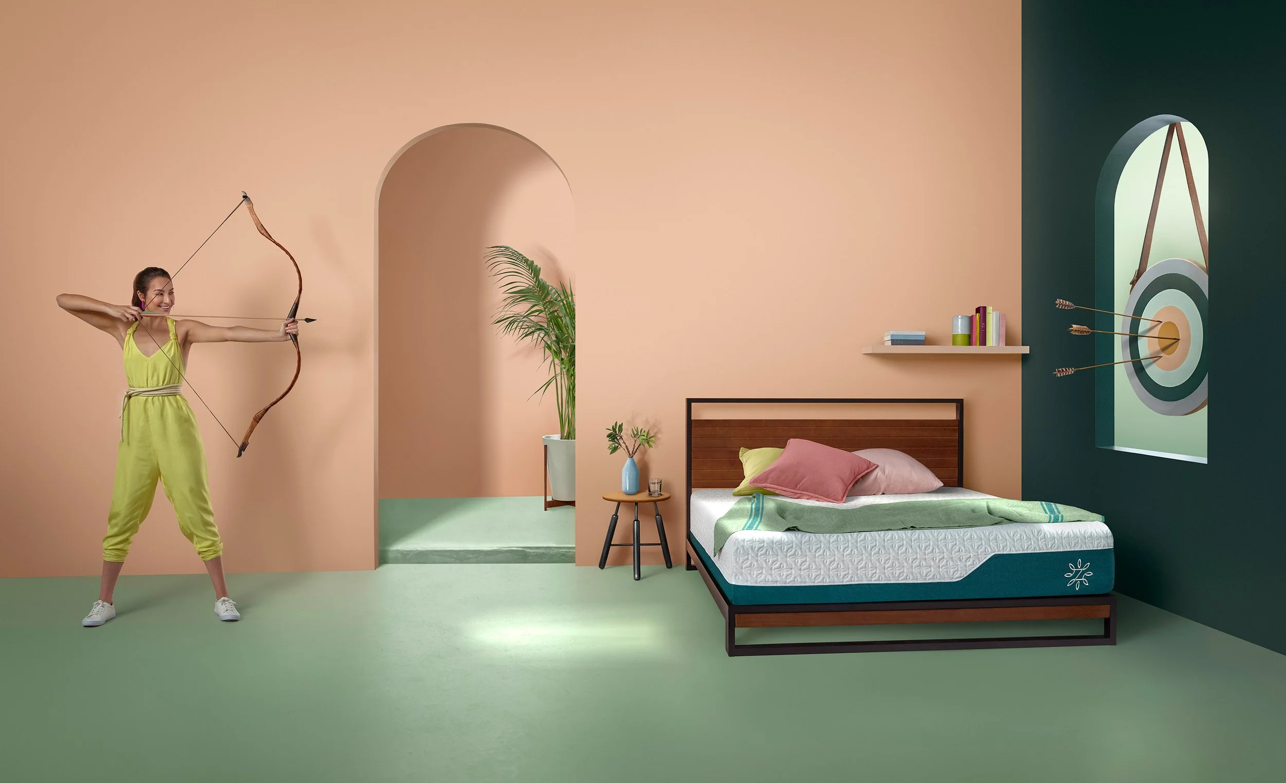 A woman dressed in a yellow outfit aims a bow and arrow in a pastel-colored room with a bed, a small side table with a plant, and a wall with a mural of an arch window with a target and arrows.
