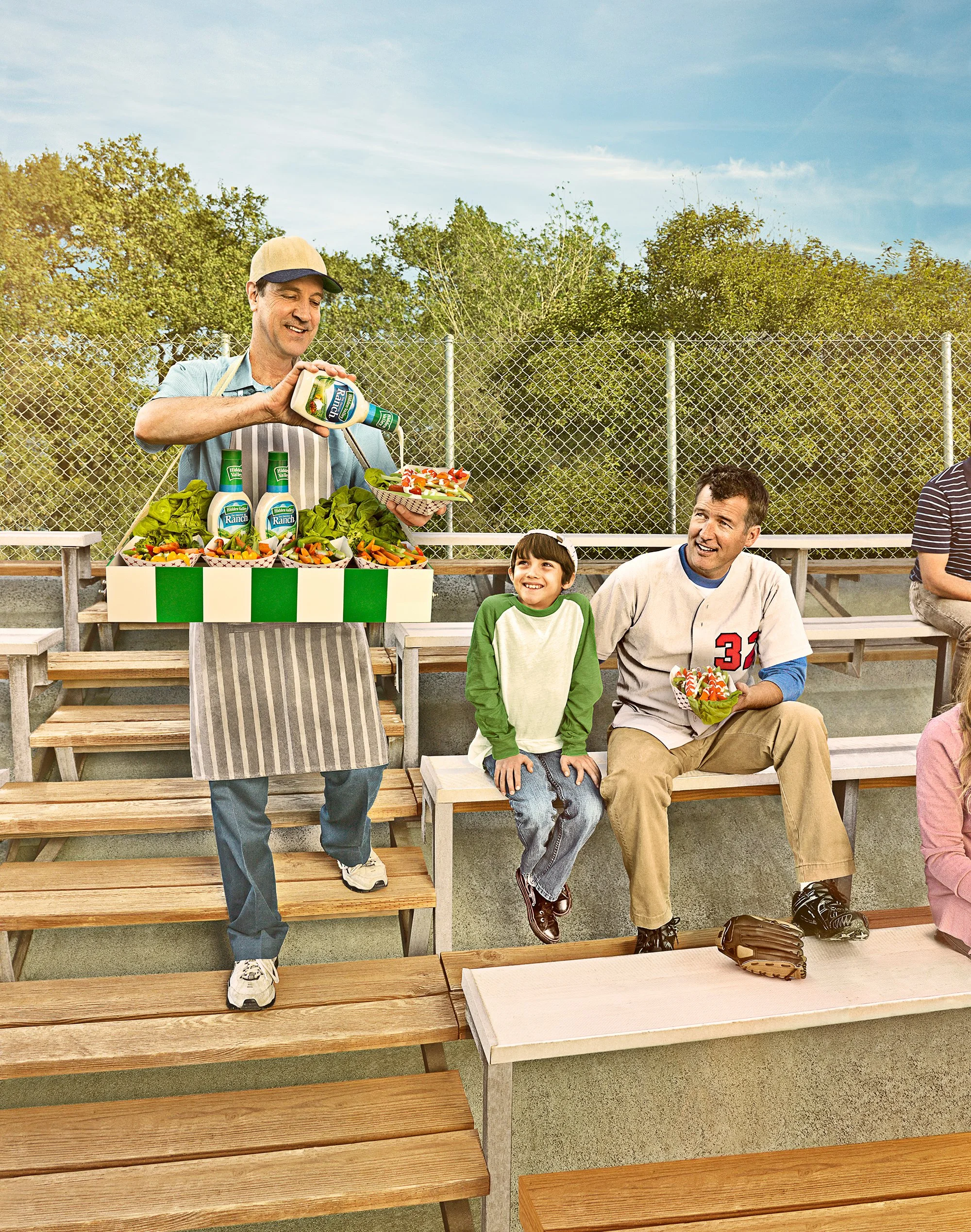 A man serving salad with Ranch dressing at a sports stadium concession stand, with a young girl and a man sitting on bleachers, a baseball glove nearby, and trees in the background.