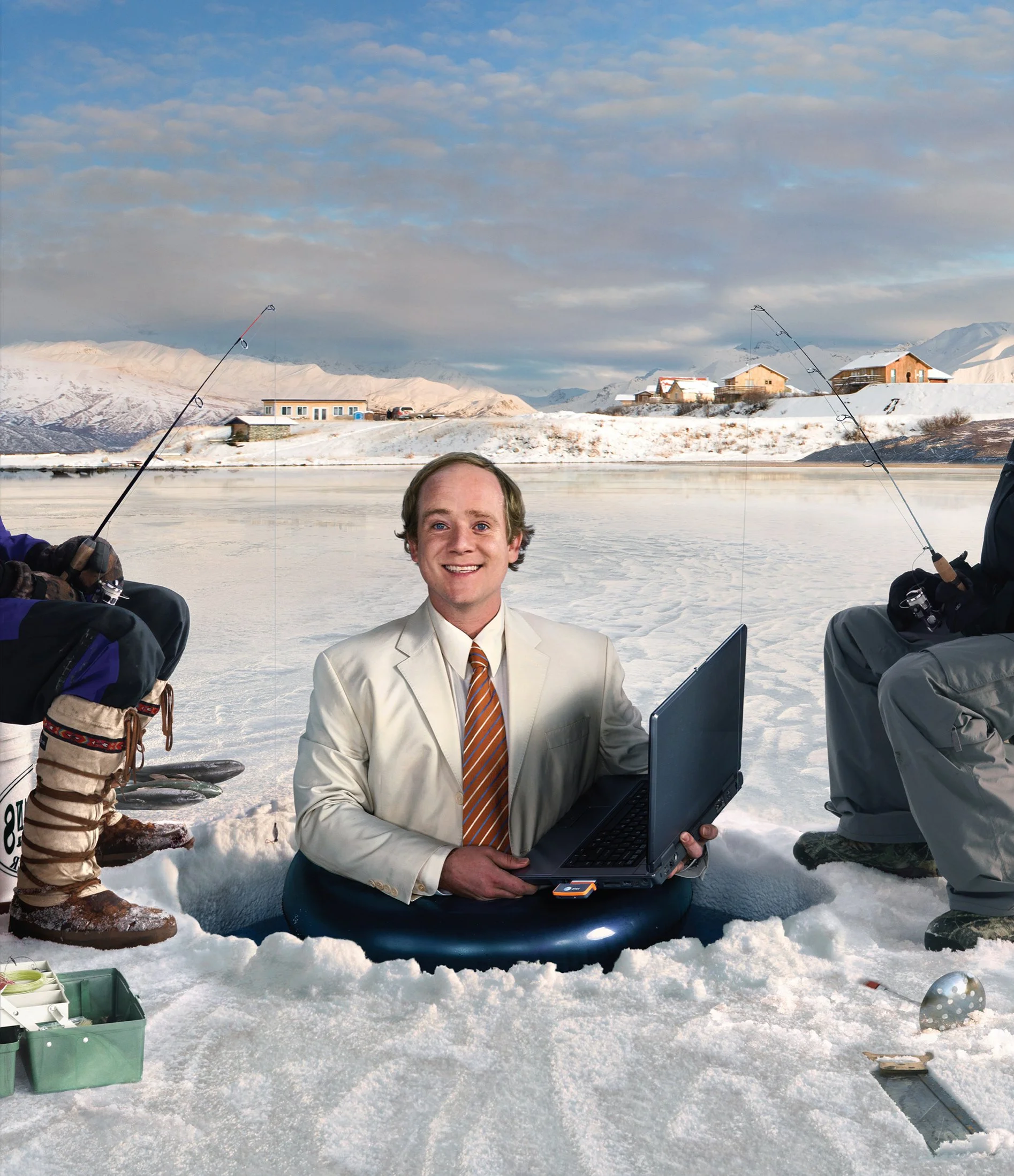 A man in a white suit with a striped tie sits in a hole in the snow, holding a laptop. Around him, people are fishing through holes in the ice on a frozen lake in a snowy landscape with houses and mountains in the background.