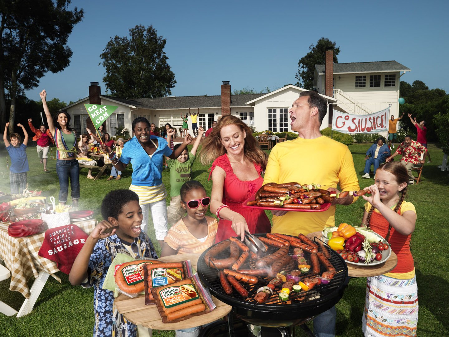 People enjoying a backyard barbecue party with grilled sausages, children and adults smiling and celebrating, with a house and party decorations in the background.