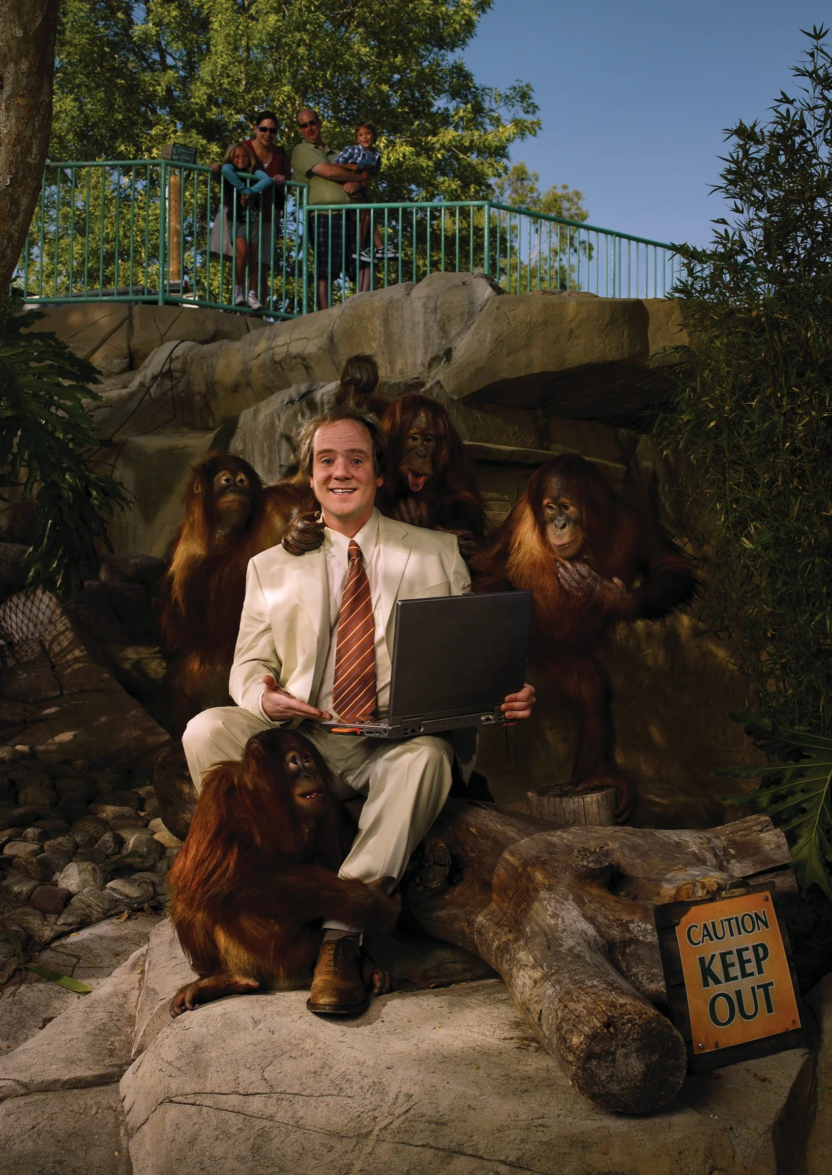 A man in a beige suit with an orange striped tie sitting on a large rock, holding a laptop, surrounded by five orangutans. Four orangutans are behind him and one sitting in front. In the background, three children and two adults stand on an elevated 