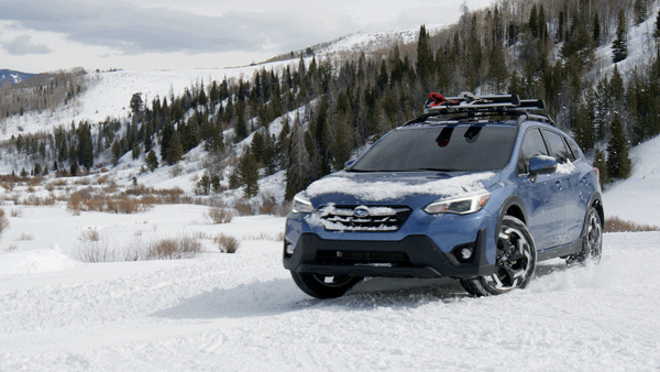 A blue Subaru Outback drives through a snowy landscape with snow-covered trees and hills in the background, equipped for winter with a roof rack.