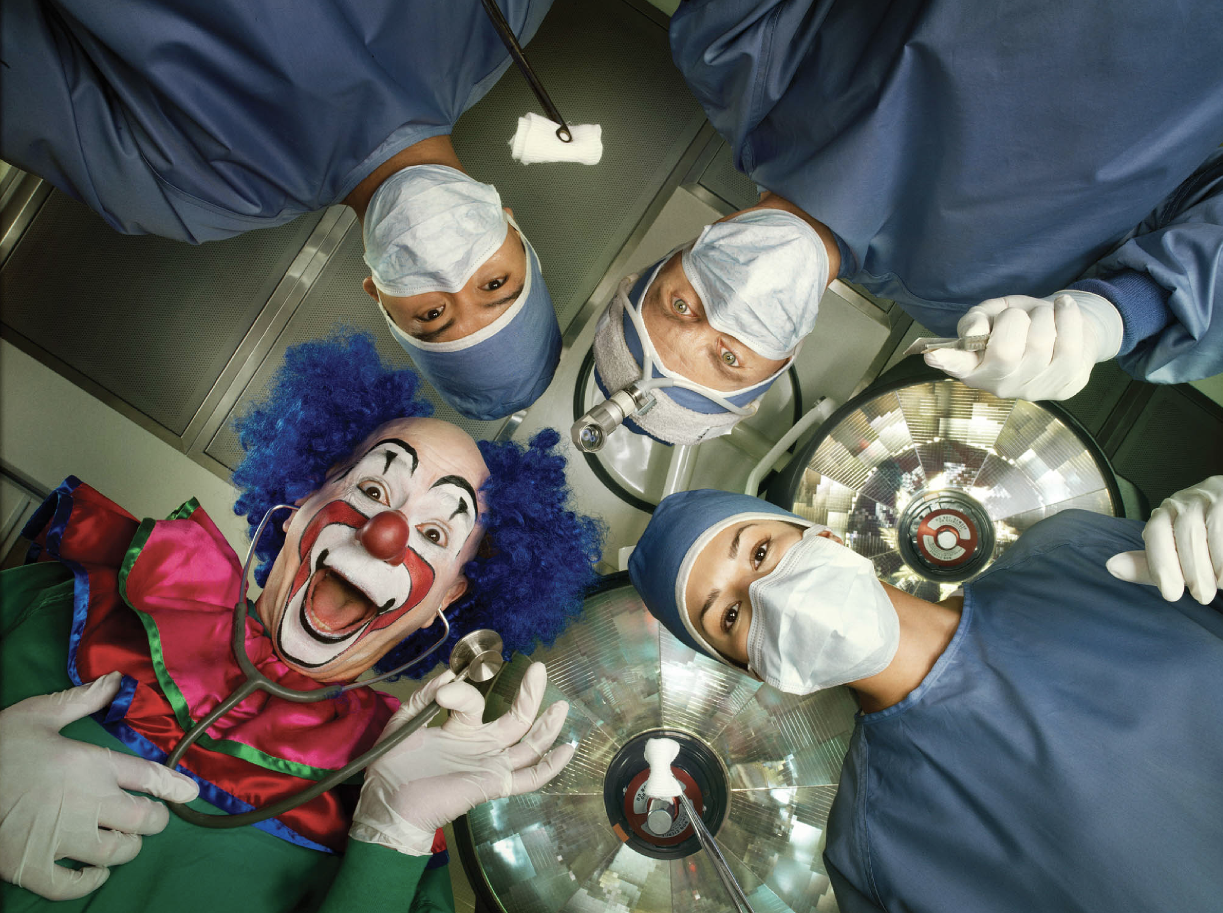 A group of five medical professionals, including a clown dressed as a clown with a red nose and colorful costume, lying on an operating table and looking down at the camera. The doctors are dressed in scrubs, surgical masks, and caps, looking up at t