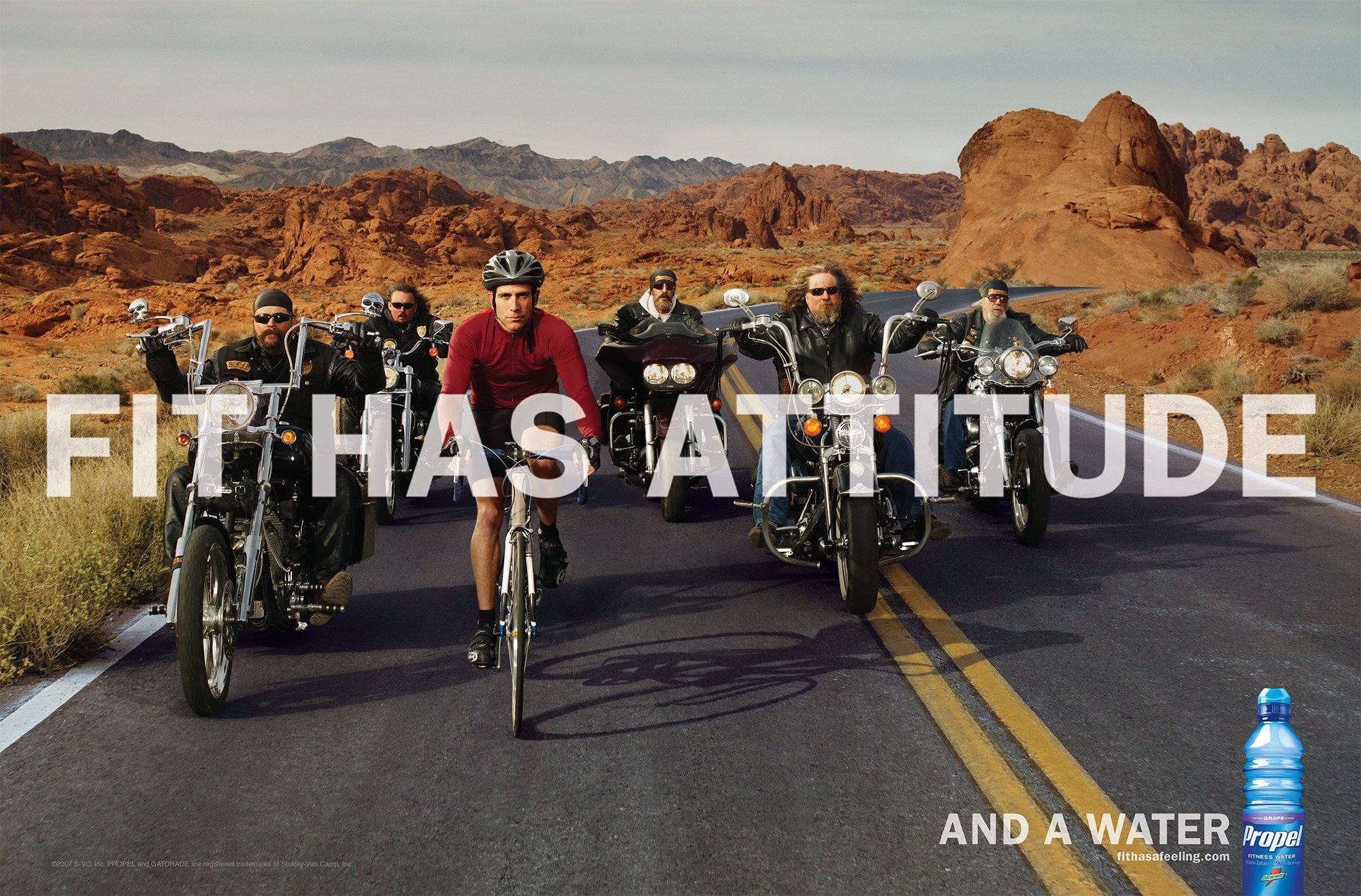 Group of six bikers on a desert road with red rock formations in the background, some on motorcycles and one on a bicycle.