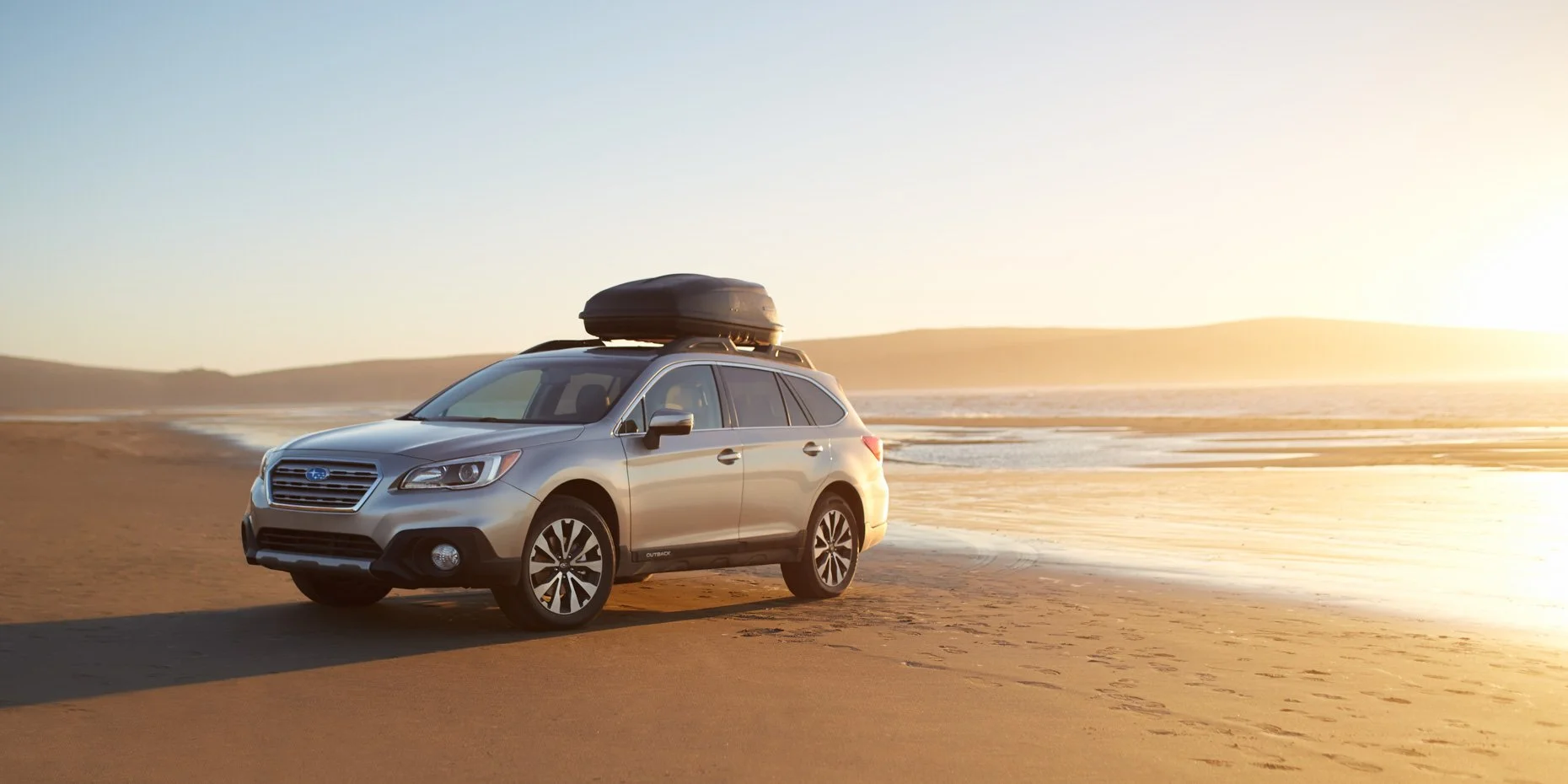 A silver Subaru Outback parked on a sandy beach at sunset with a cargo box on the roof, overlooking the ocean and distant hills.