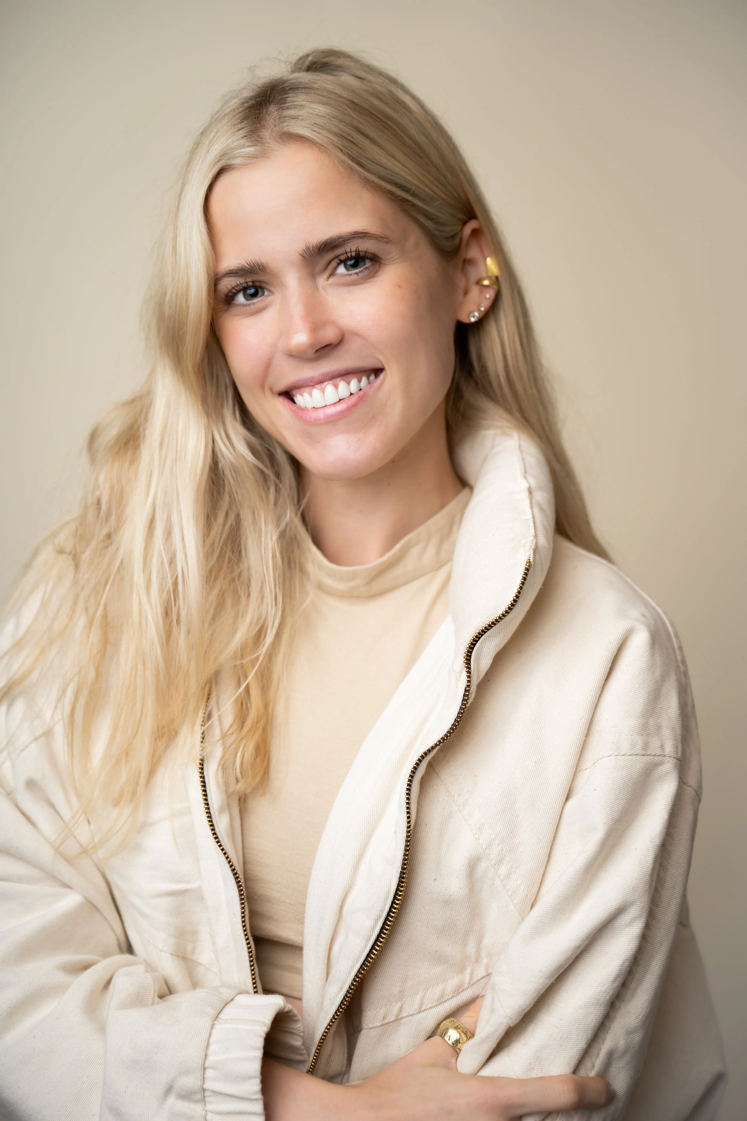 A young woman with long blonde hair, smiling, wearing a beige zip-up jacket and earrings, posing against a plain background.