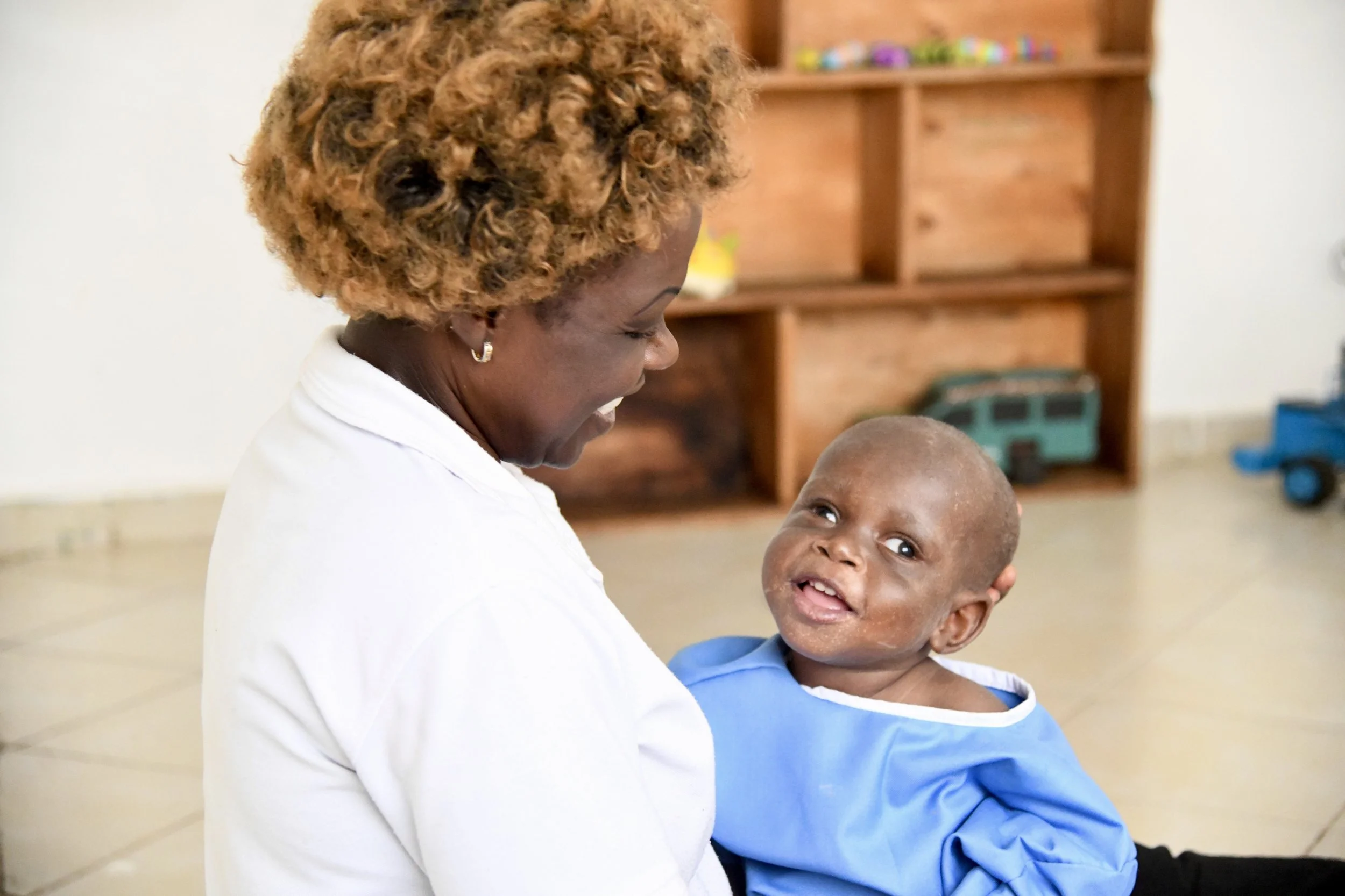 A woman with curly, reddish-brown hair and wearing a white shirt is smiling at a young boy. The boy has a shaved head, is wearing a blue shirt, and has some visible facial scars. They are indoors, with a wooden shelf and some toy vehicles in the background.