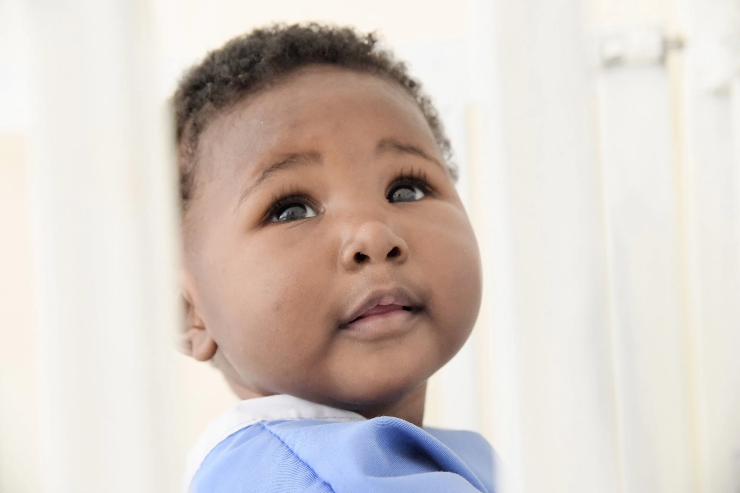 A young child with short curly hair and dark eyes looking upwards, wearing a light blue smock or shirt. The background is light-colored with vertical slats.