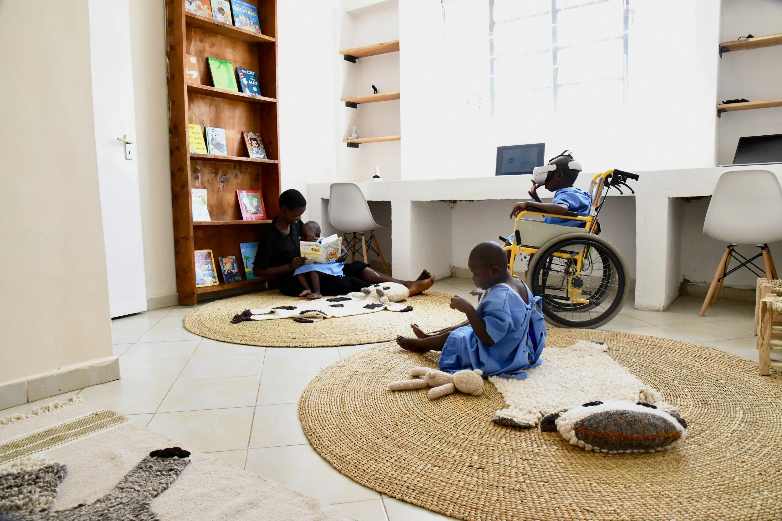 A room with three children, two sitting on woven rugs and one in a wheelchair, engaging in different activities. One child is reading a book to a younger one, another is sitting and eating, and the third in a wheelchair is using a virtual reality headset. The room has a wooden bookshelf with children's books, a white tiled floor, and a large window.