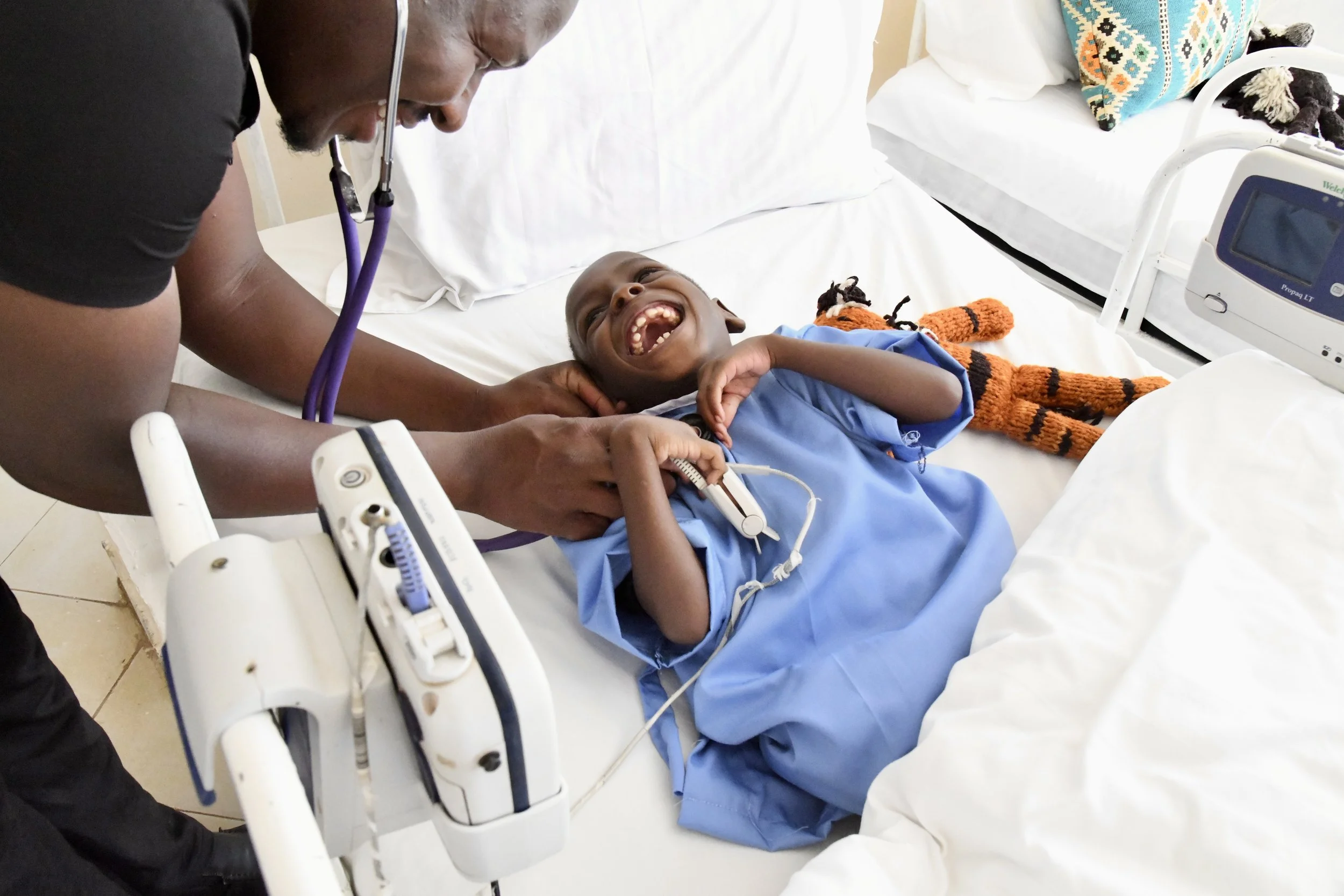 A smiling young boy lying in a hospital bed, being examined by a healthcare professional with a stethoscope.