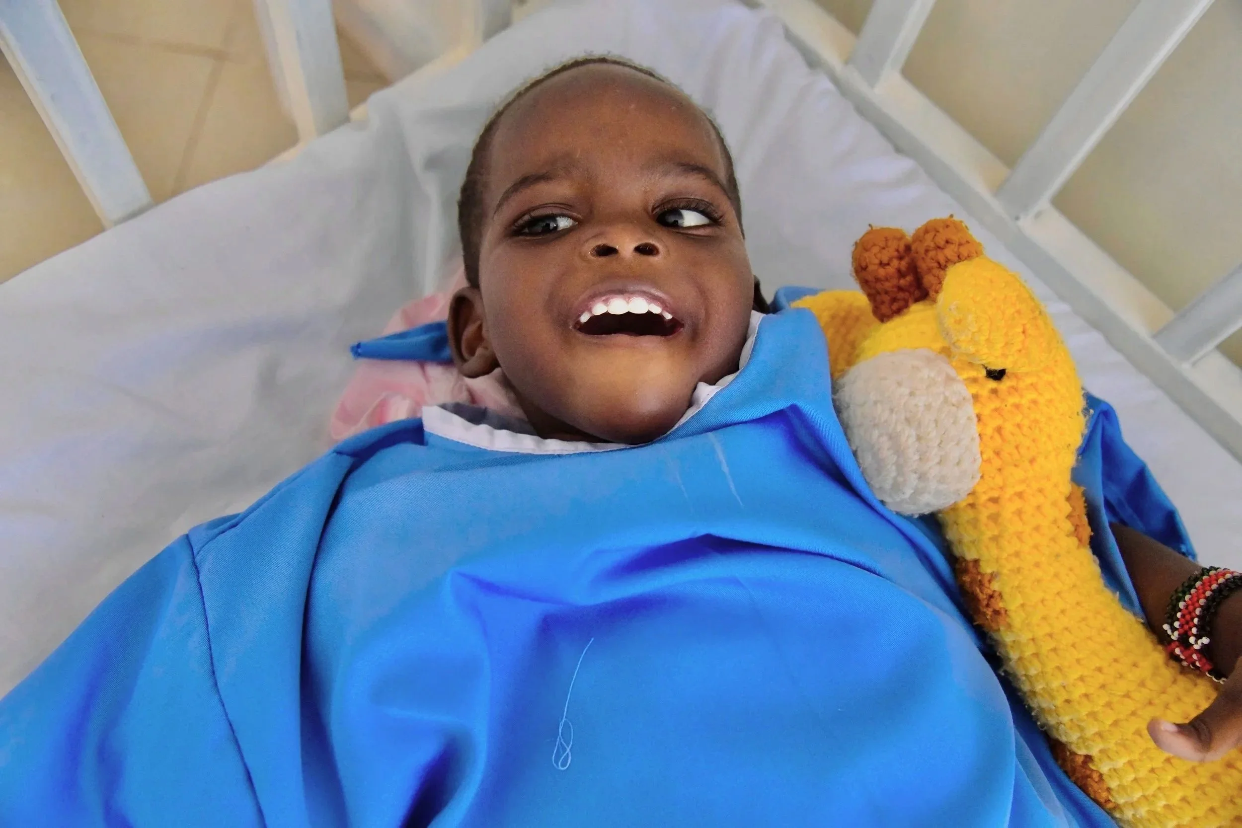 A young child lying in a hospital bed, smiling, holding a crocheted giraffe plush toy, wearing a blue hospital gown.