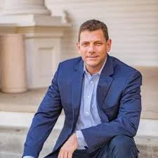 A man in a blue suit sitting outdoors in front of a white building with columns.