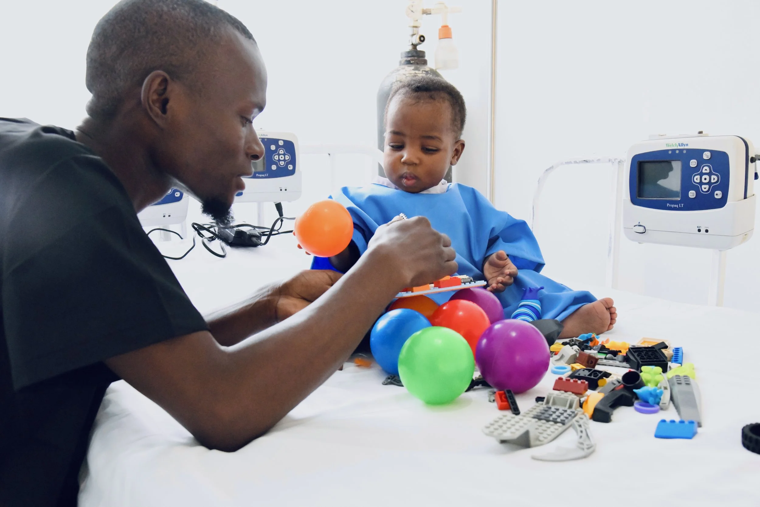 A young boy in a hospital bed playing with colorful toys and a man assisting him.