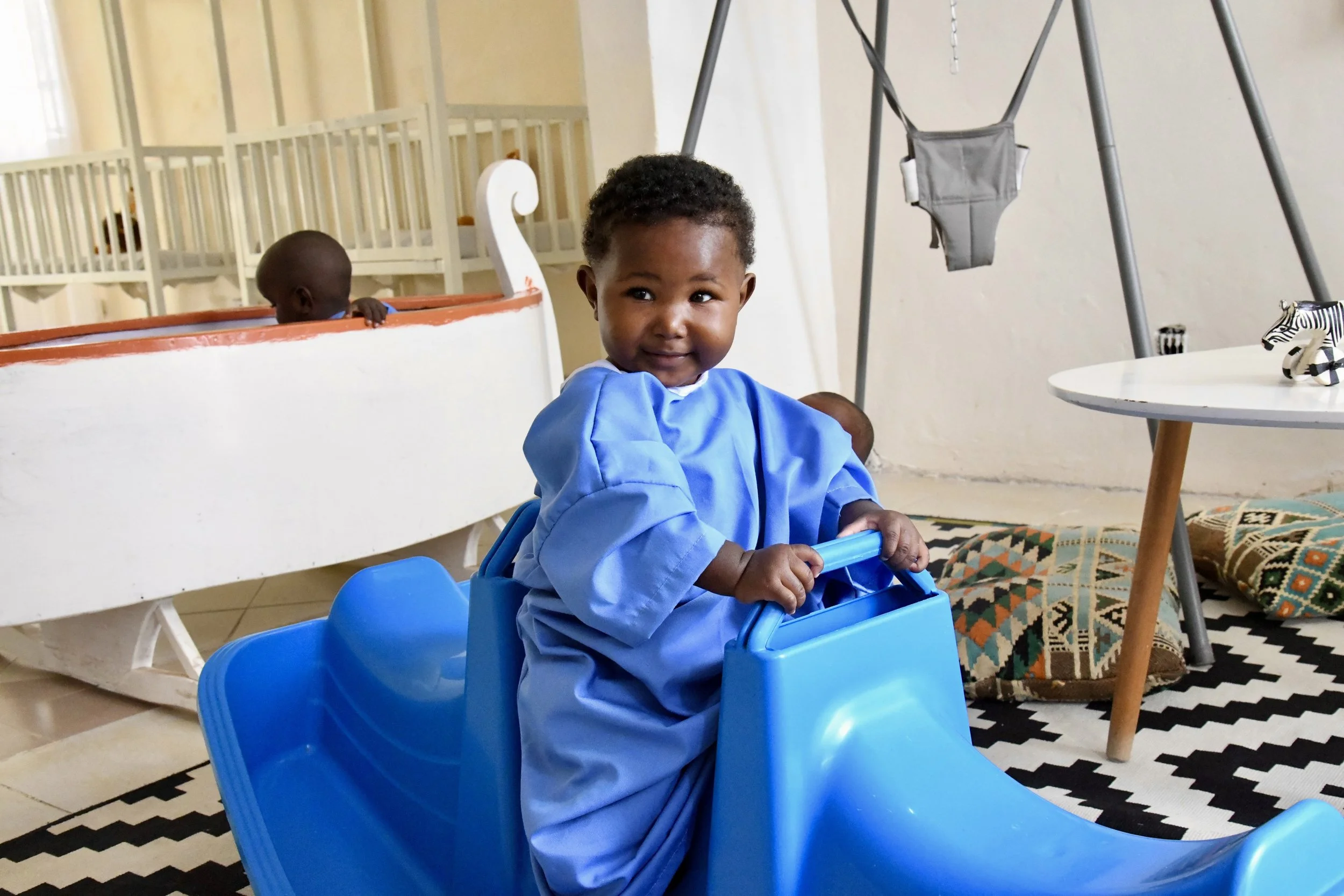 A young child in a blue smock smiling while sitting on a blue plastic slide in a playroom with a black and white patterned rug, a round white table, and a pillow in the background.
