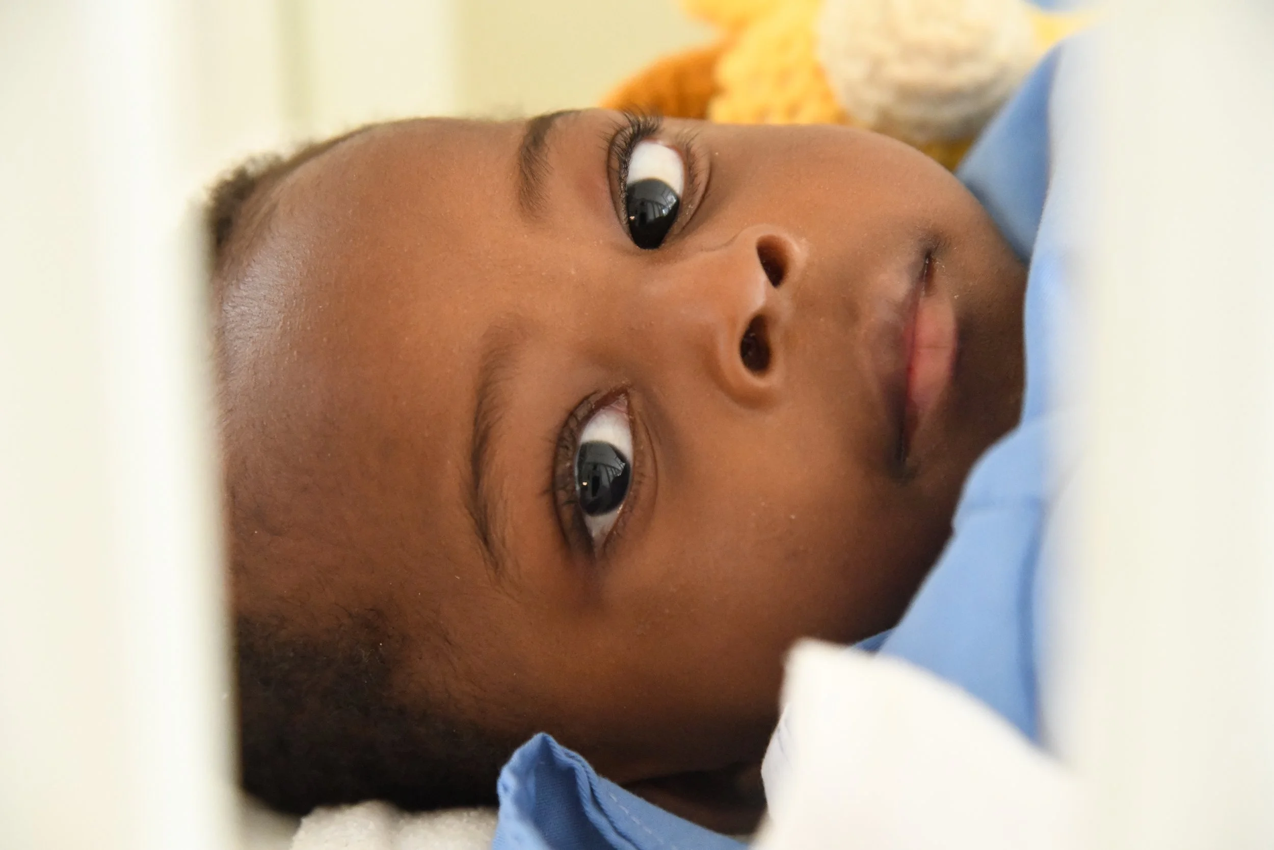Close-up of a young boy lying down, looking up with a gentle expression, wearing a light blue shirt.