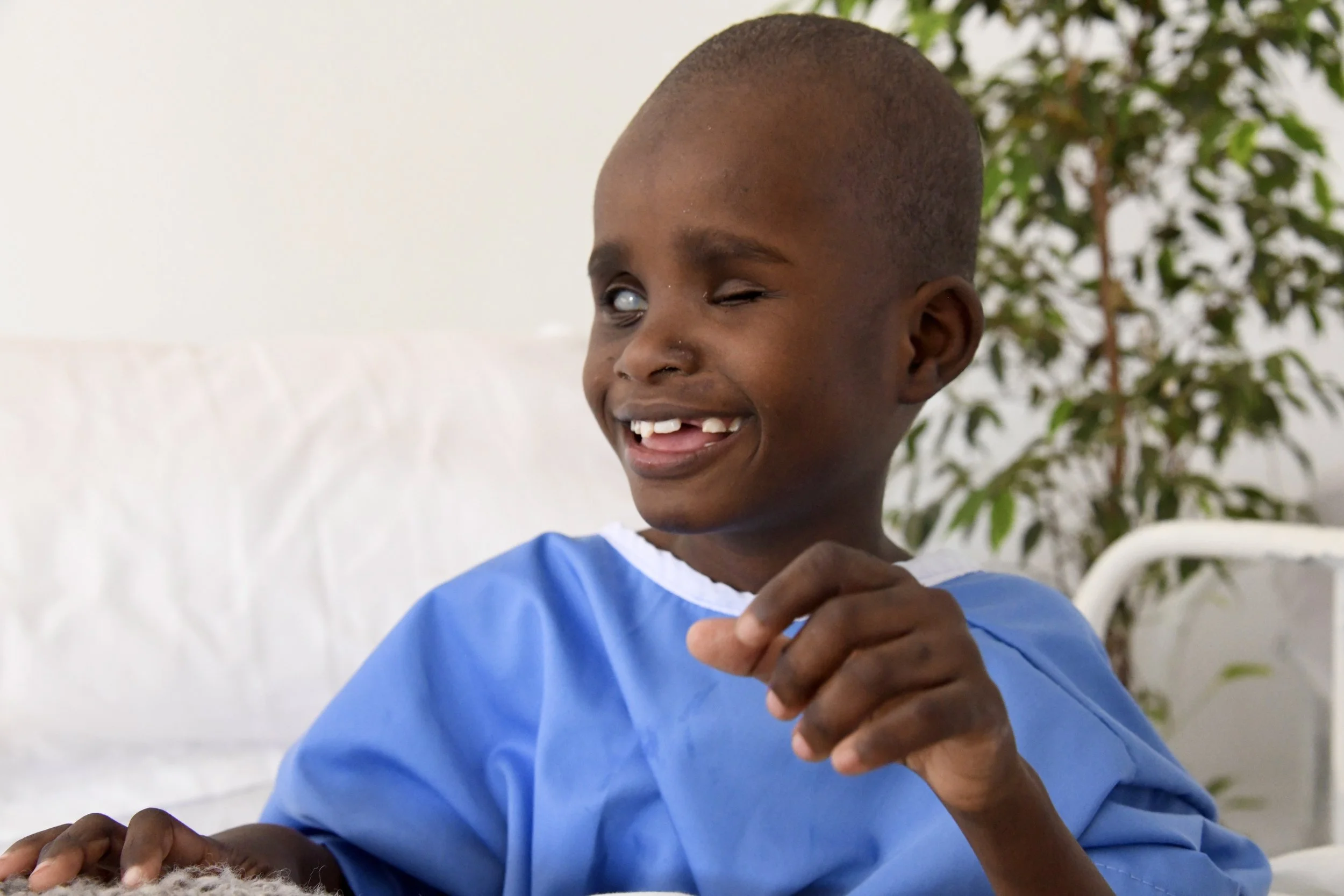 A young boy with three missing front teeth smiling and winking while looking to the side, wearing a blue hospital gown, sitting in a room with a white bed and a potted plant in the background.