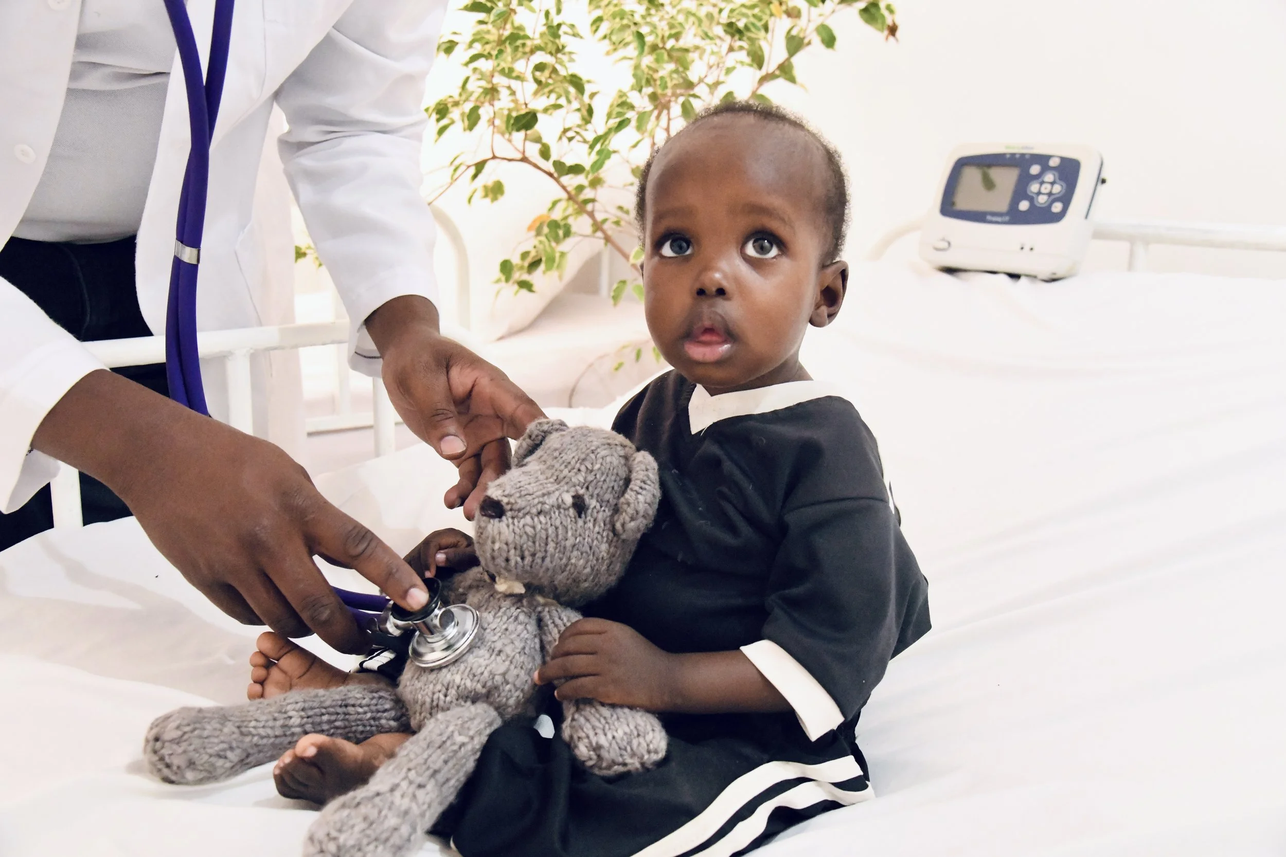 A young child sitting on a hospital bed, being examined by a doctor. The child is holding a knitted teddy bear and has a surprised or curious expression. The doctor is wearing a white coat and stethoscope, and is using a stethoscope on the child's teddy bear. In the background, there is a medical monitor and some greenery.