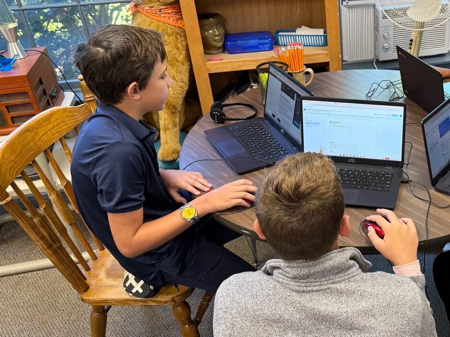Two boys working on laptops at a round table with various electronic devices in a room with wooden furniture and a window.