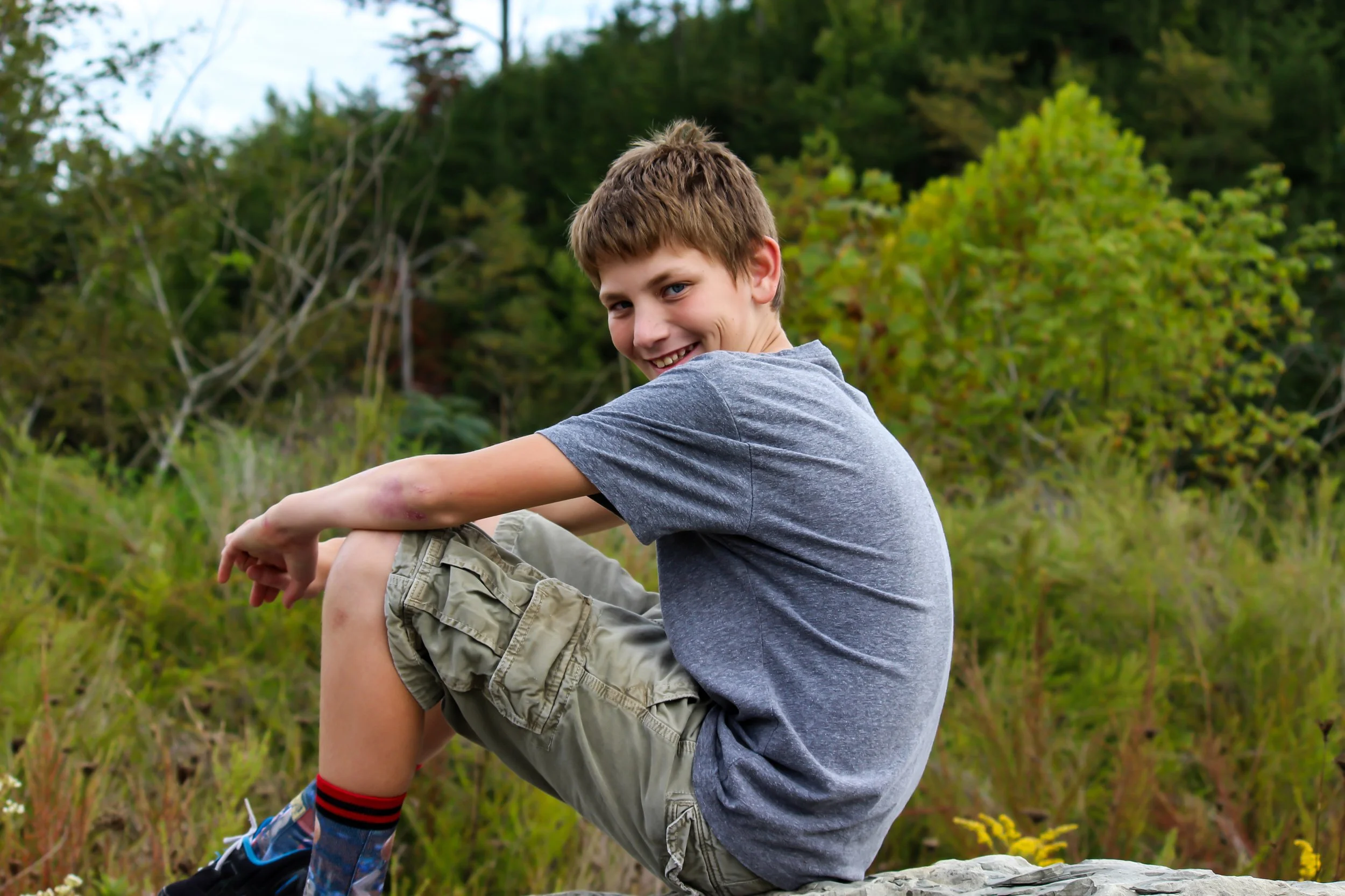 A young boy sitting outdoors on a rock, smiling and looking back at the camera, surrounded by green trees and vegetation.
