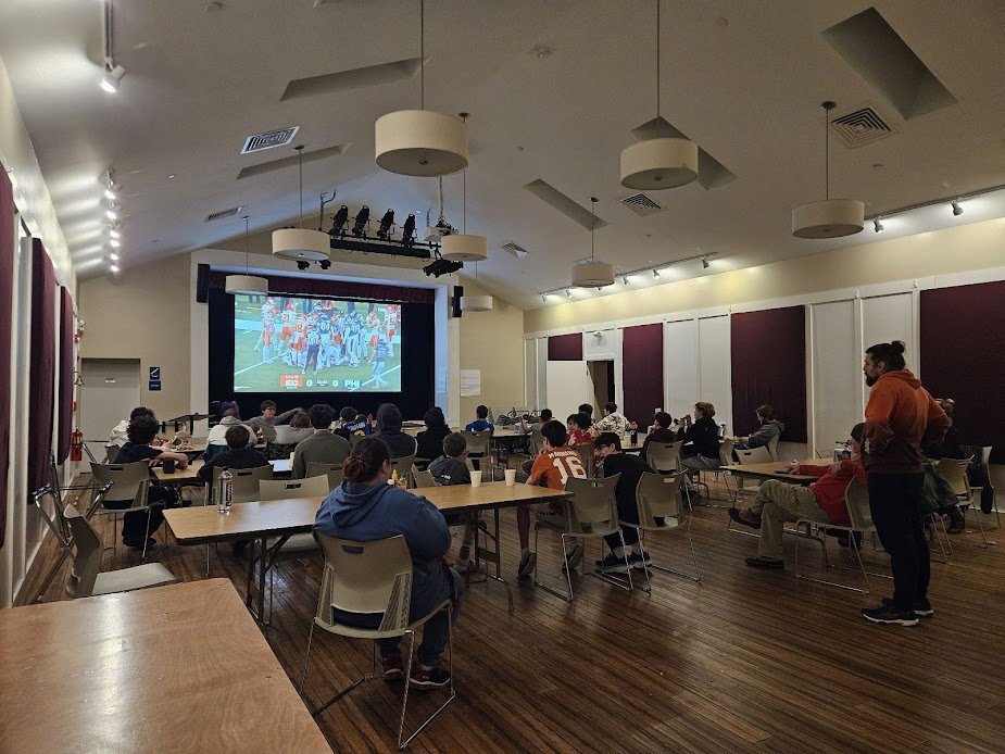 People watching a sports game on a large screen in a spacious room with wooden floors, ceiling lights, and purple curtains.