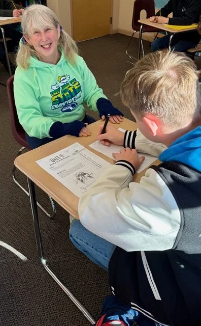 A young woman with long blonde hair in pigtails, wearing a bright green hoodie and blue gloves, smiling while sitting at a desk with a young boy. The boy, with blond hair, is sitting across from her, wearing a white jacket with black stripes and holding a pen over a worksheet.