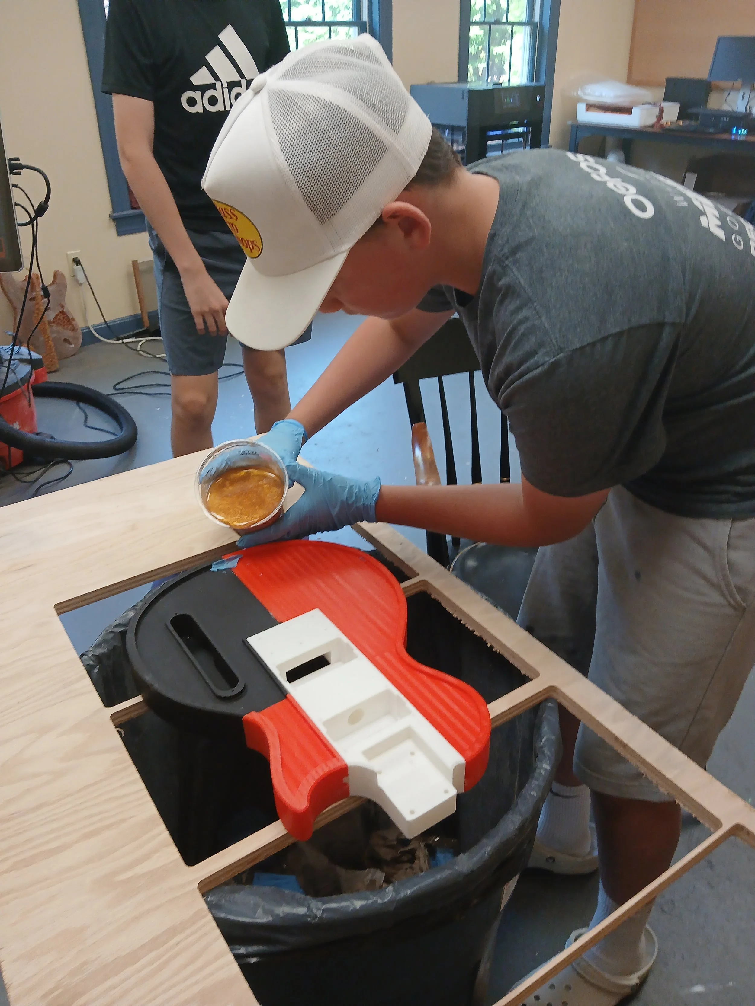 A man wearing a white cap and gray t-shirt working on a woodworking project, applying a liquid to a piece of wood, with another person standing nearby in a workshop.