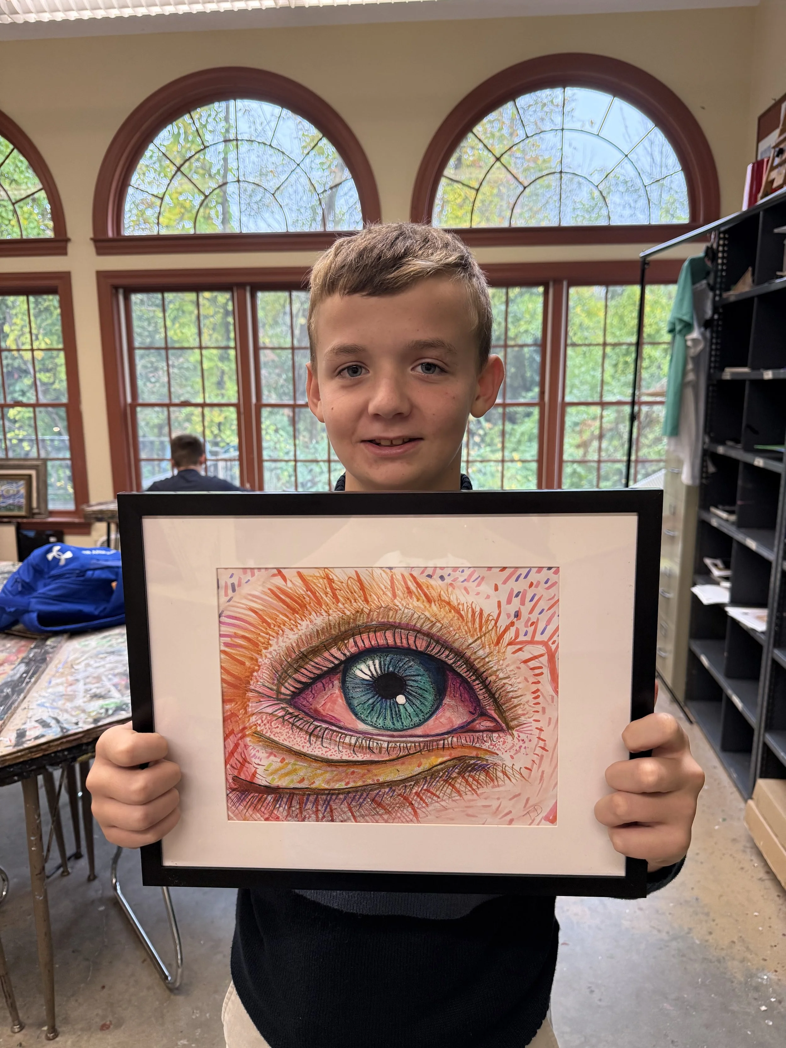 A young boy holding a framed colorful drawing of a human eye in an art classroom with large arched windows and a view of trees outside.