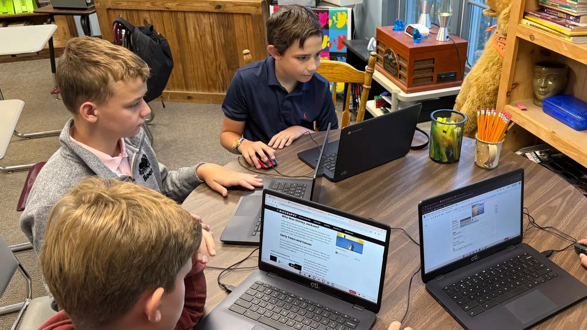 Four children sitting at a wooden table working on laptops in a classroom or library. One boy in a navy shirt is using a mouse, children appear focused.