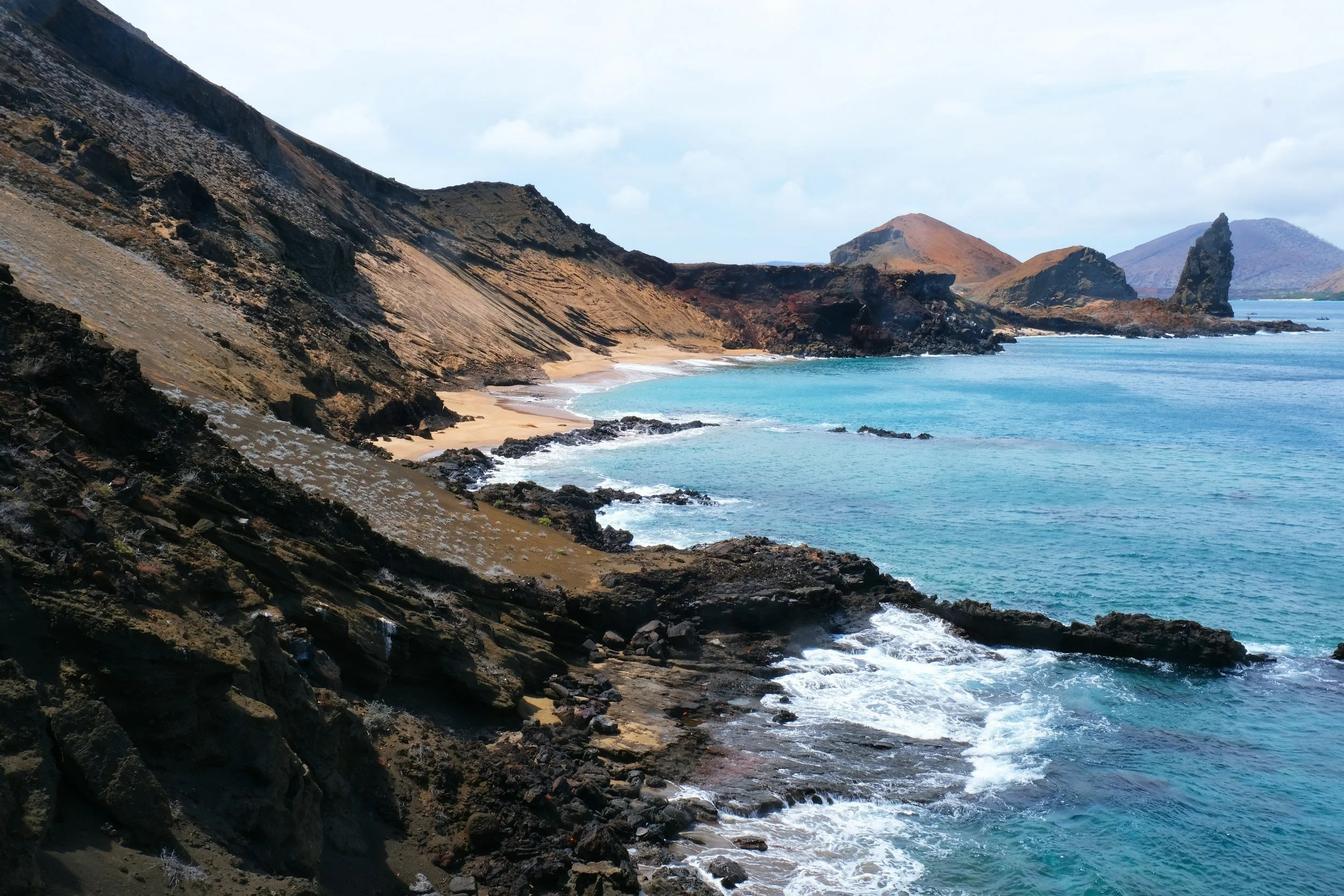 Coastal landscape with rocky cliffs, sandy beach, and turquoise ocean waters, with distant hills and cloudy sky.