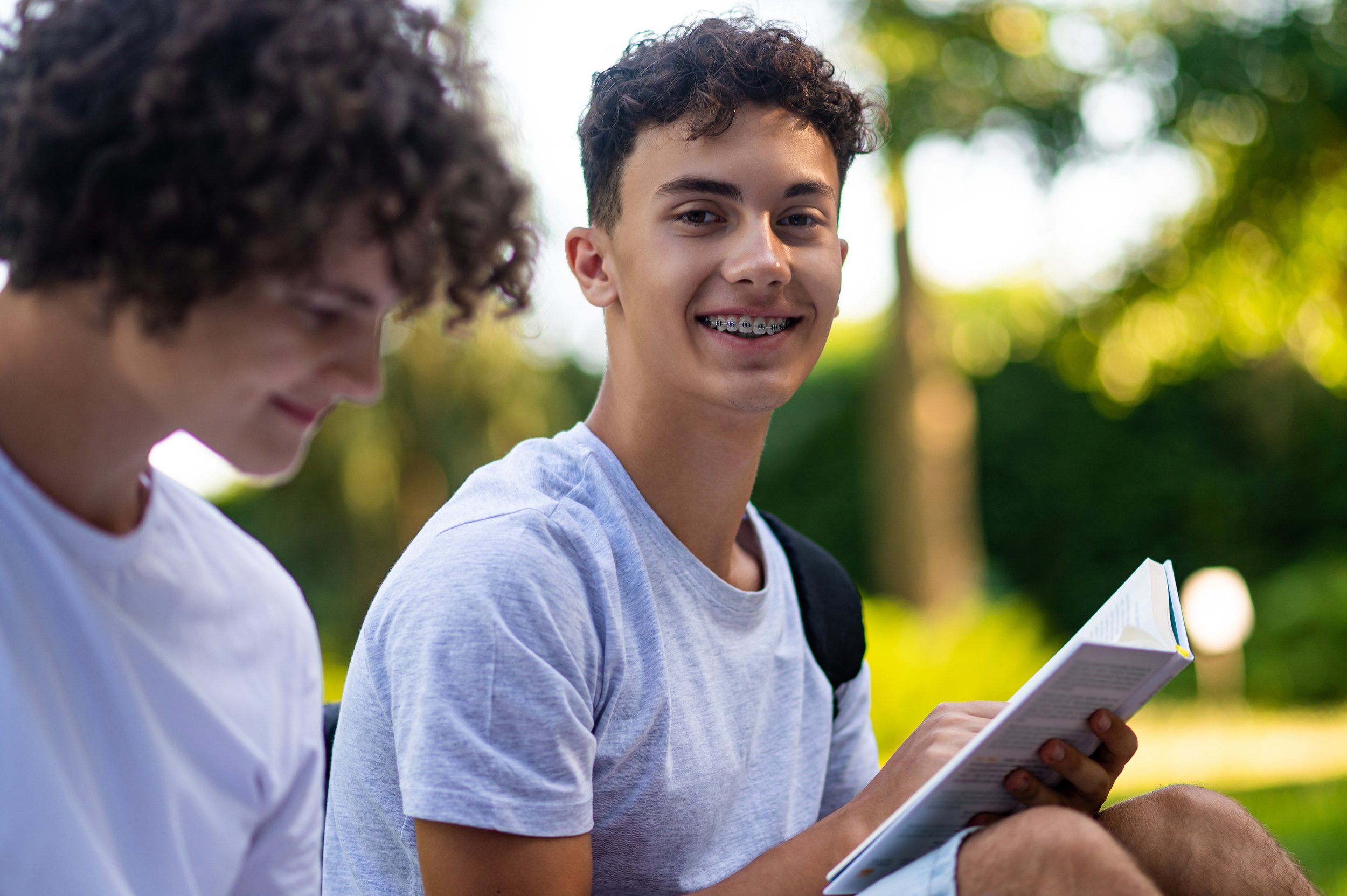 Two teenage boys sitting outdoors in a park, one is smiling at the camera holding a book, and the other is looking down, with trees and greenery in the background.