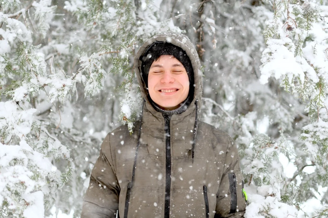 A young man smiling with eyes closed, wearing a brown winter jacket and a black beanie, standing amidst snow-covered trees during a snowfall.