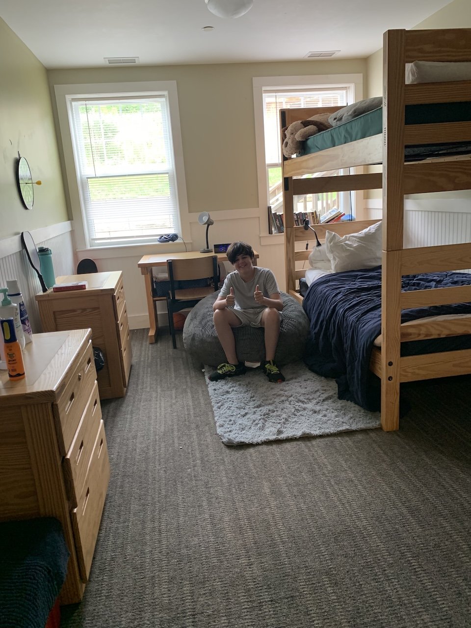 A boy sitting on a gray bean bag chair in a bedroom, giving a thumbs-up. The room has two large windows, bunk beds, a wooden dresser, and a small desk with a chair. The walls are painted light green and white, and there is a small white rug on the floor.