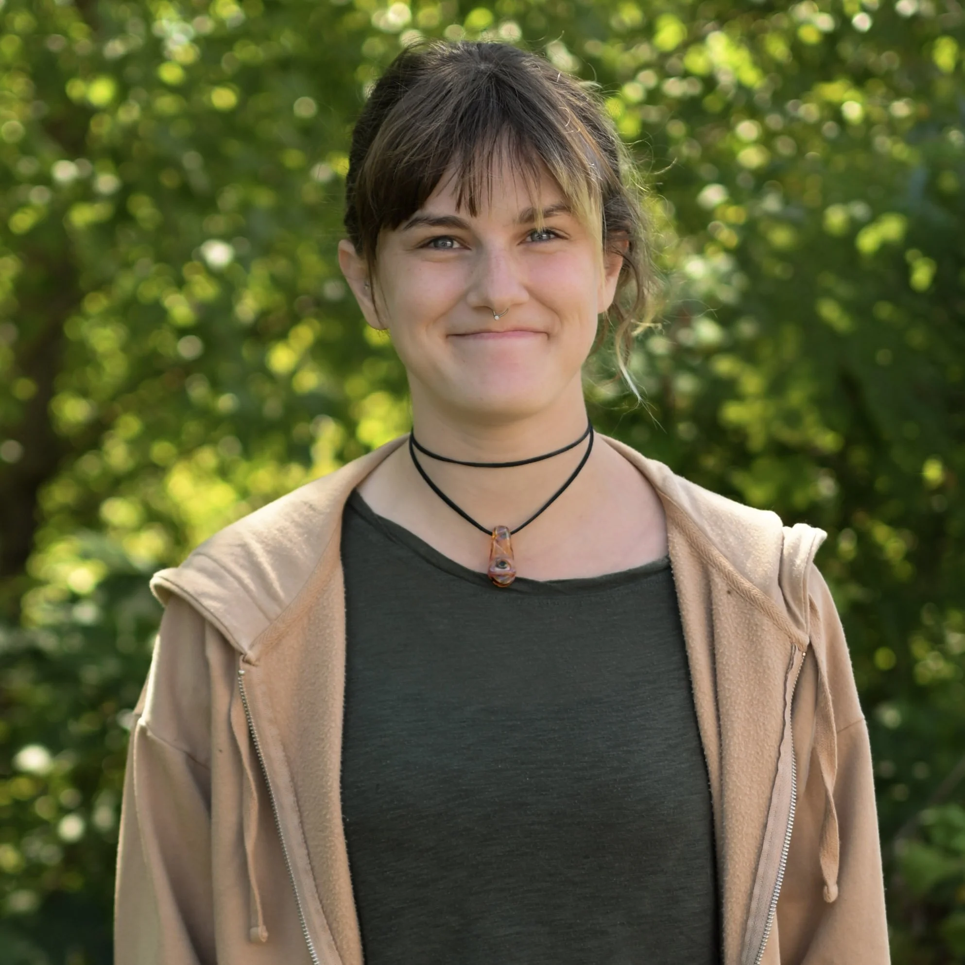 A young woman with short brown hair, wearing a black T-shirt, beige hoodie, and layered necklaces, standing outdoors with green trees in the background.