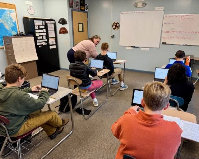 A classroom with six students sitting at desks working on laptops, with a teacher assisting one student. The classroom has a whiteboard, educational posters, and a map on the walls.