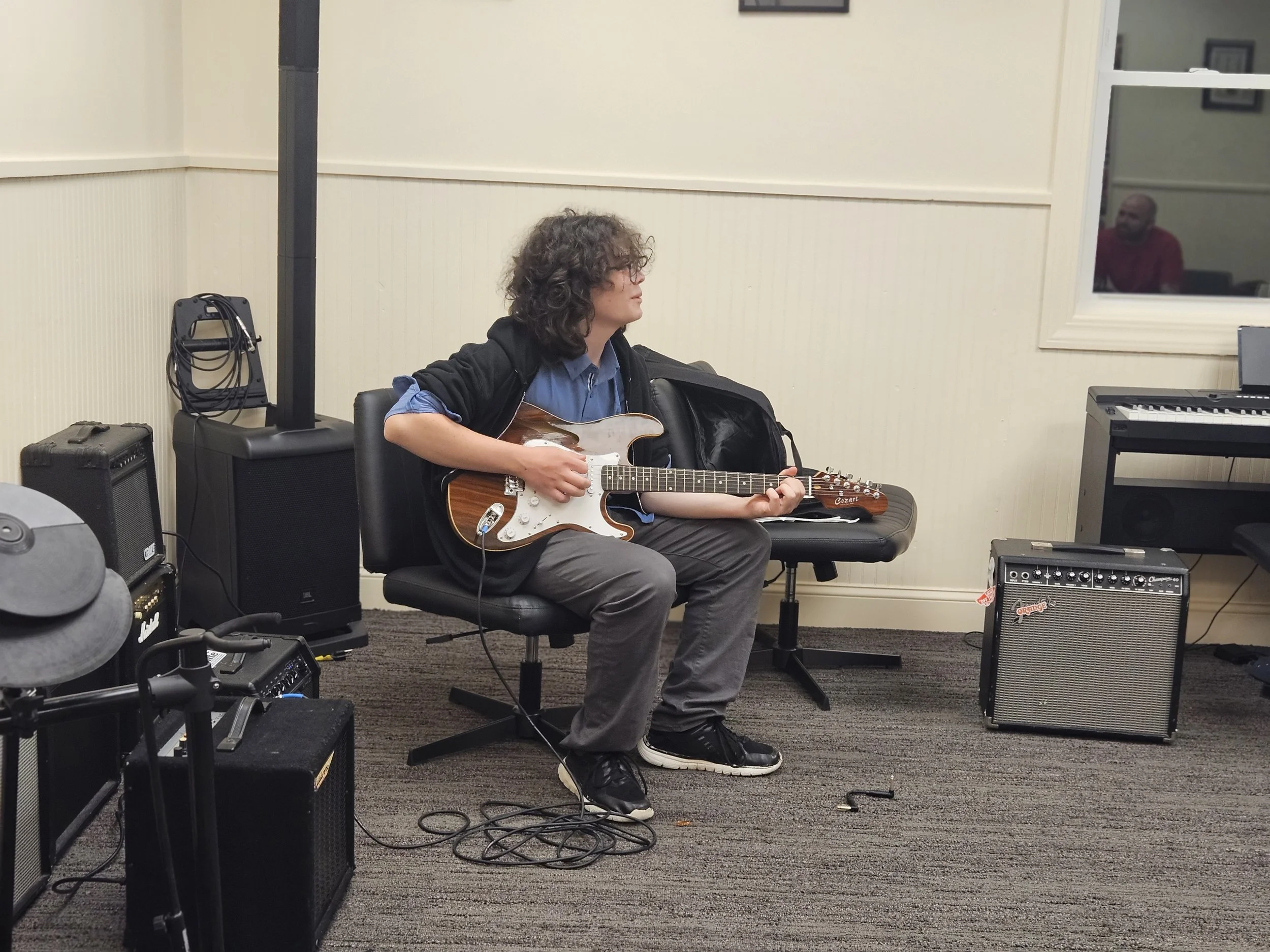A person with curly hair sitting on a black chair, playing an electric guitar in a music rehearsal space surrounded by amplifiers and musical equipment.