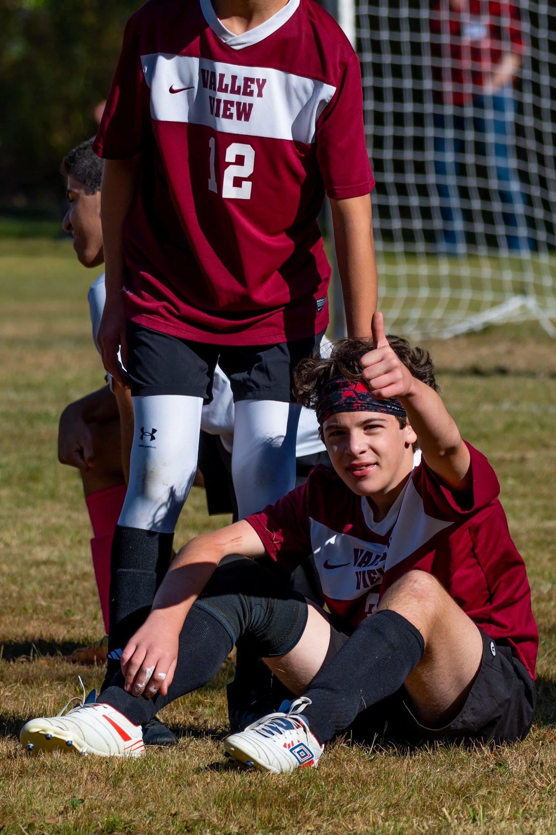 A group of young soccer players on a field, with one sitting on the ground giving a thumbs-up gesture. The players are wearing maroon and white jerseys, and there is a soccer goal in the background.