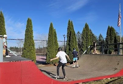 People skateboarding at an outdoor skatepark with tall trees and a blue sky in the background.