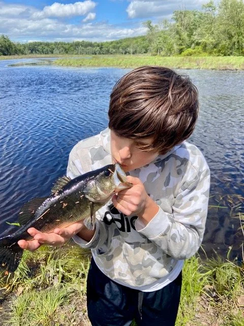 A young boy with brown hair holding a fish he caught near a riverbank, with lush green trees and a partly cloudy sky in the background.