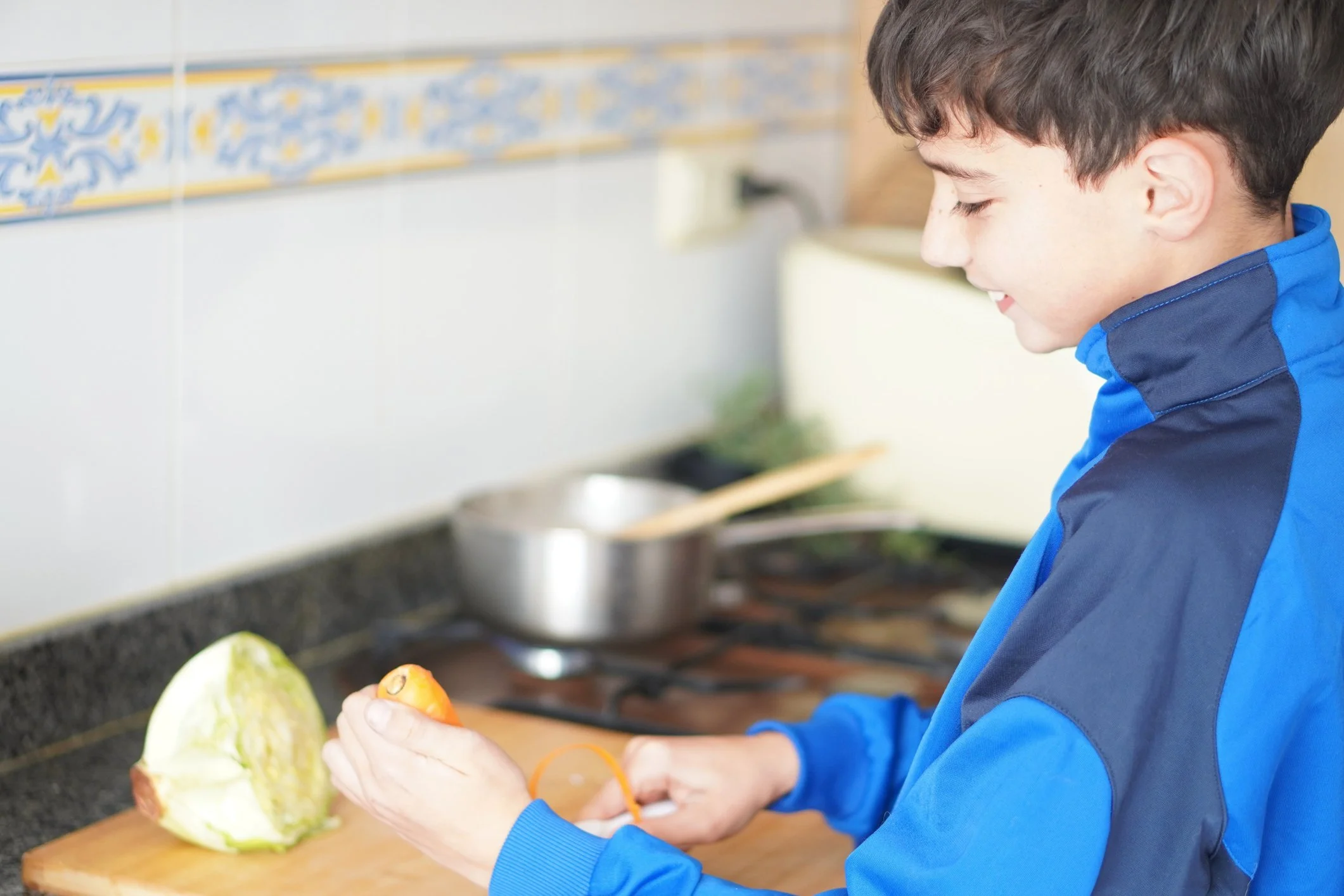 A boy peeling an orange in a kitchen, with a head of lettuce on a cutting board nearby.