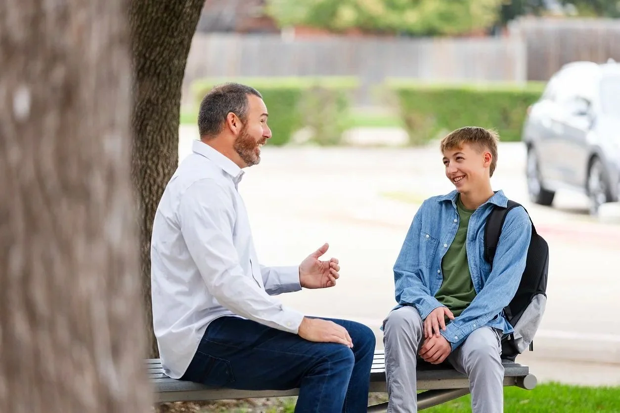 A man and a boy sitting on a park bench, engaged in conversation, with a tree trunk partially blocking the view.