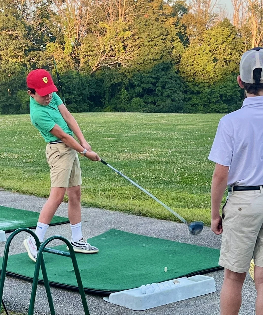 A boy wearing a red cap and green shirt practicing golf at a driving range, with another boy in a white shirt and khaki shorts watching nearby.
