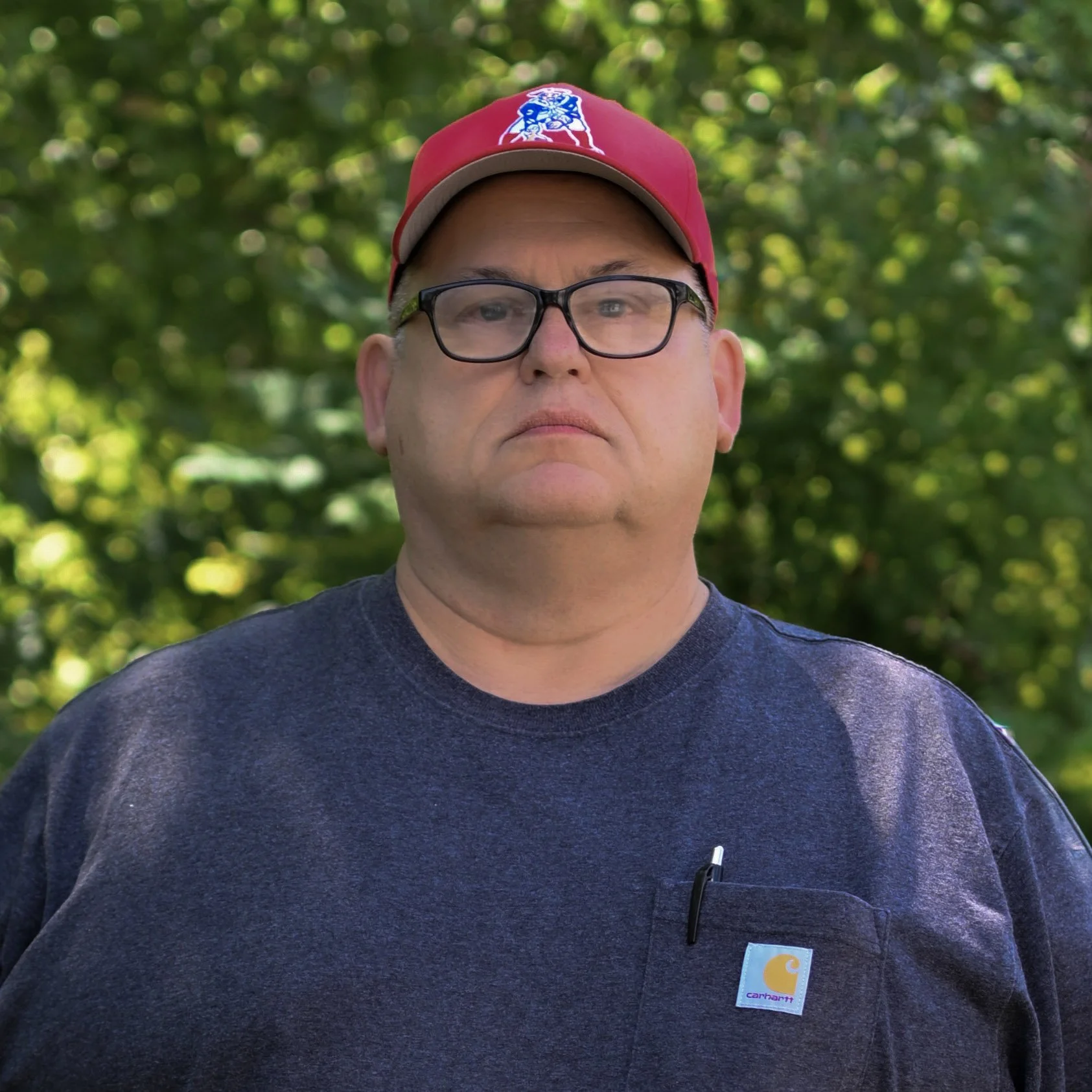 A man wearing glasses, a red baseball cap with a sports logo, and a dark t-shirt with a small pocket on the chest and a Carhartt logo, standing outdoors with a background of green foliage.