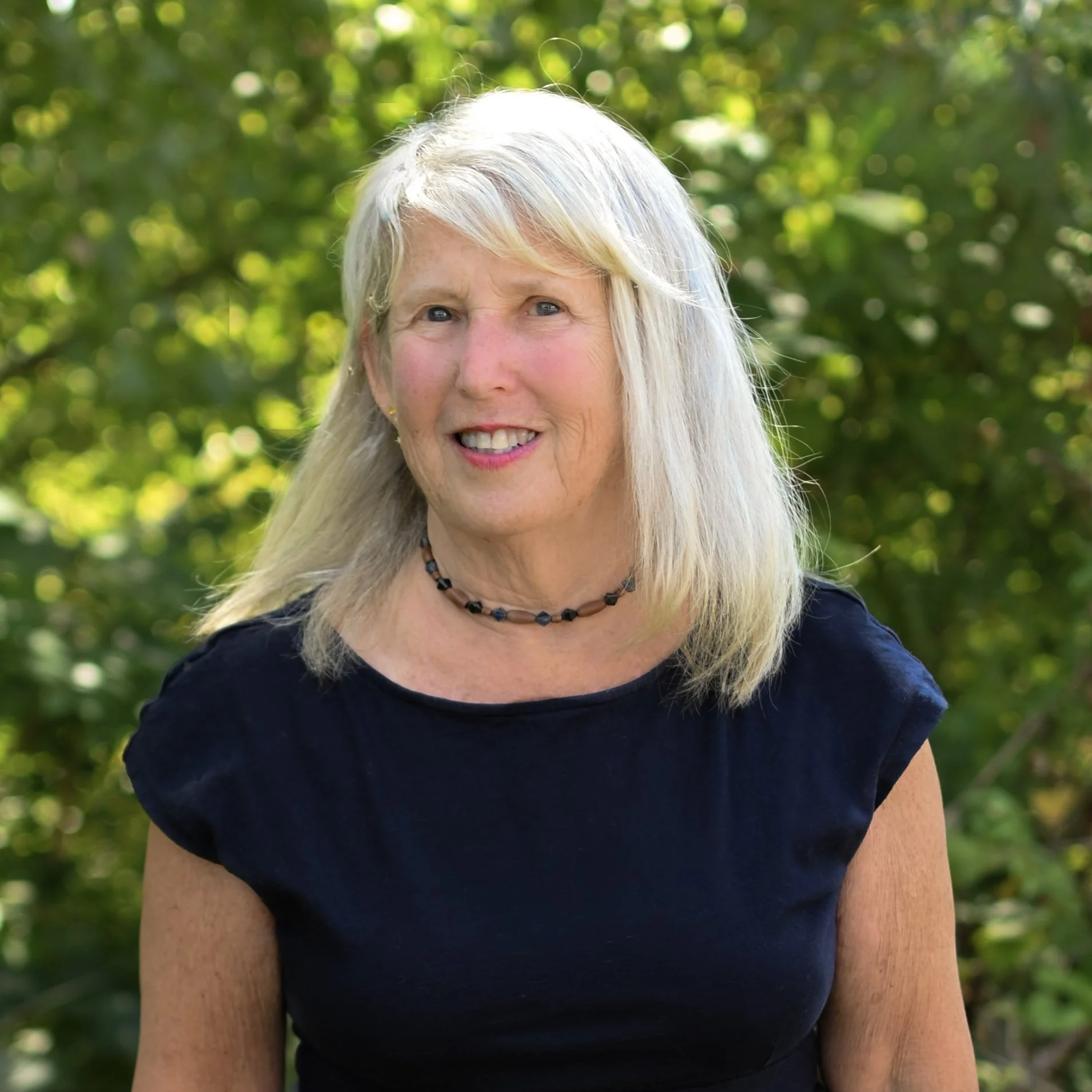 A smiling woman with blonde hair and blue eyes wearing a black top and a black necklace, outdoors with green foliage in the background.