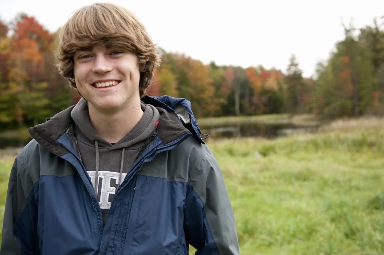 Young man smiling outdoors with autumn trees and a pond in the background.
