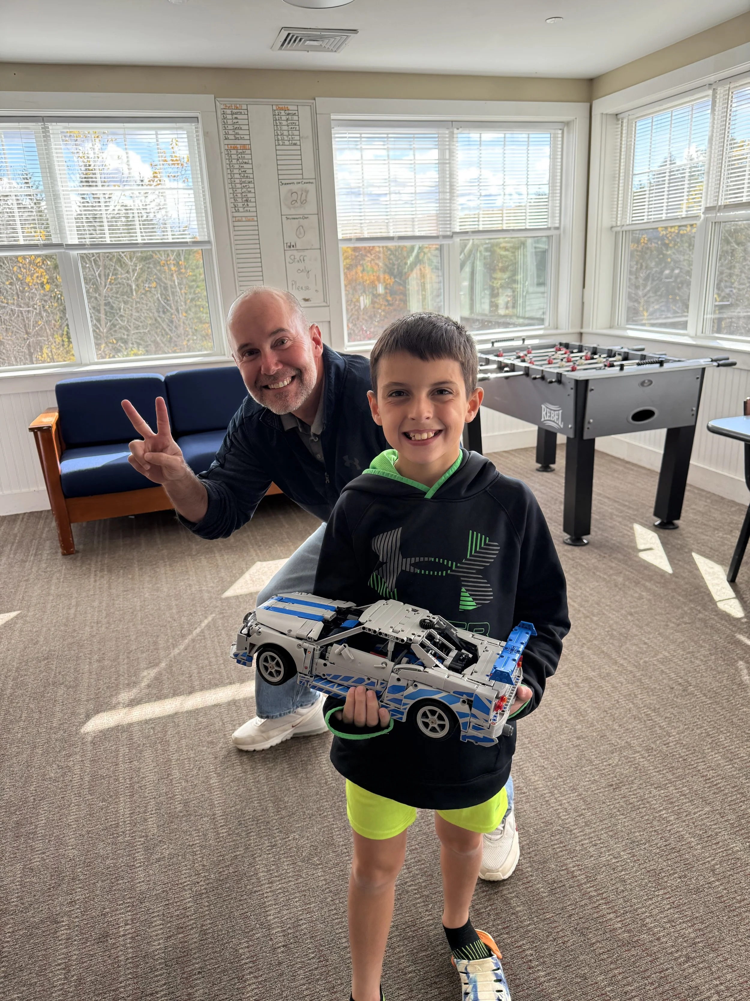 A smiling young boy in a black hoodie and bright green shorts holding a LEGO car model, standing in a sunlit room with windows and a foosball table. A man behind him is smiling and giving a peace sign.