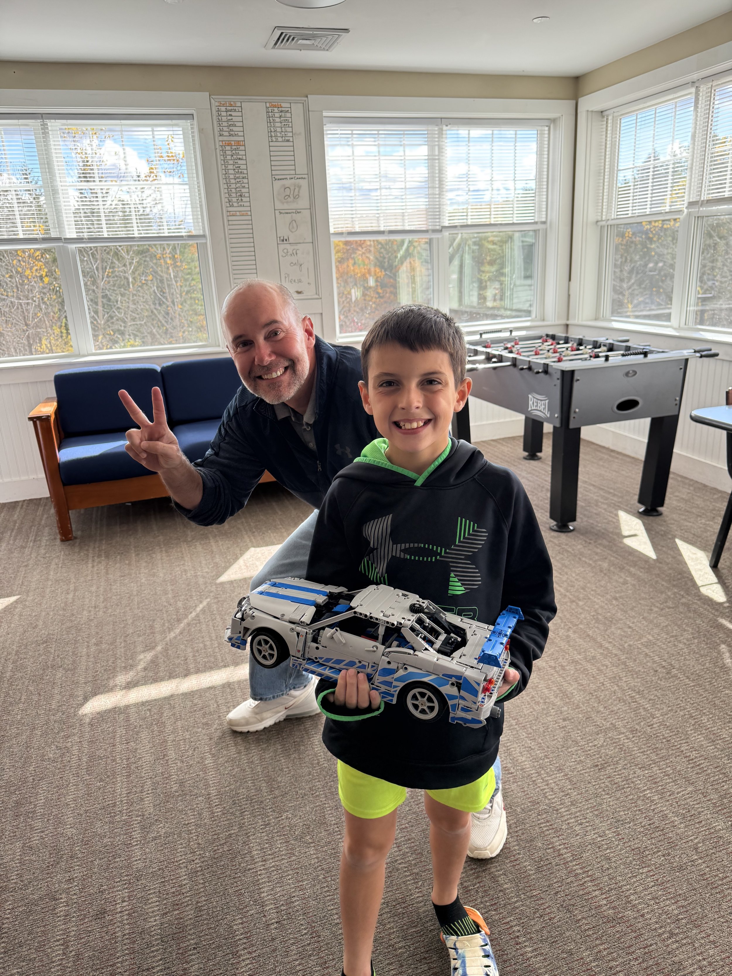 A smiling young boy in a black hoodie and bright green shorts holding a LEGO car model, standing in a sunlit room with windows and a foosball table. A man behind him is smiling and giving a peace sign.